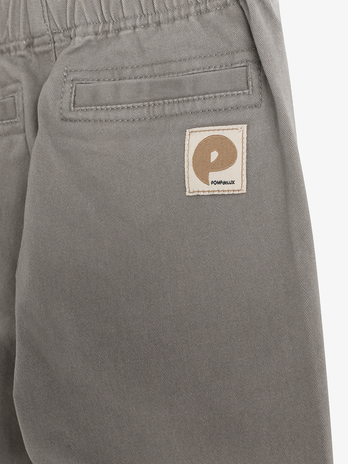Gray fabric pants feature a patch with a stylized letter "P" and the text "POMPdeLUX" near the pocket, against a plain background.