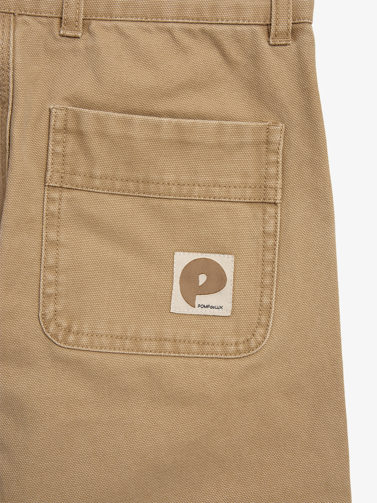 Beige fabric pocket on pants with a logo patch showing a stylized "P" and text "POMPdeLUX" on a square label, against the same material background.
