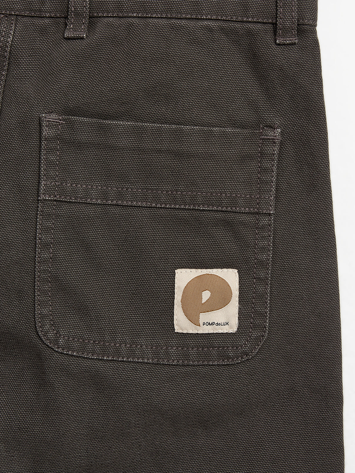 Dark fabric pants pocket featuring a beige patch with a stylized "P" logo. Text reads "POMPdeLUX." Belt loops visible at the top in a plain background setting.