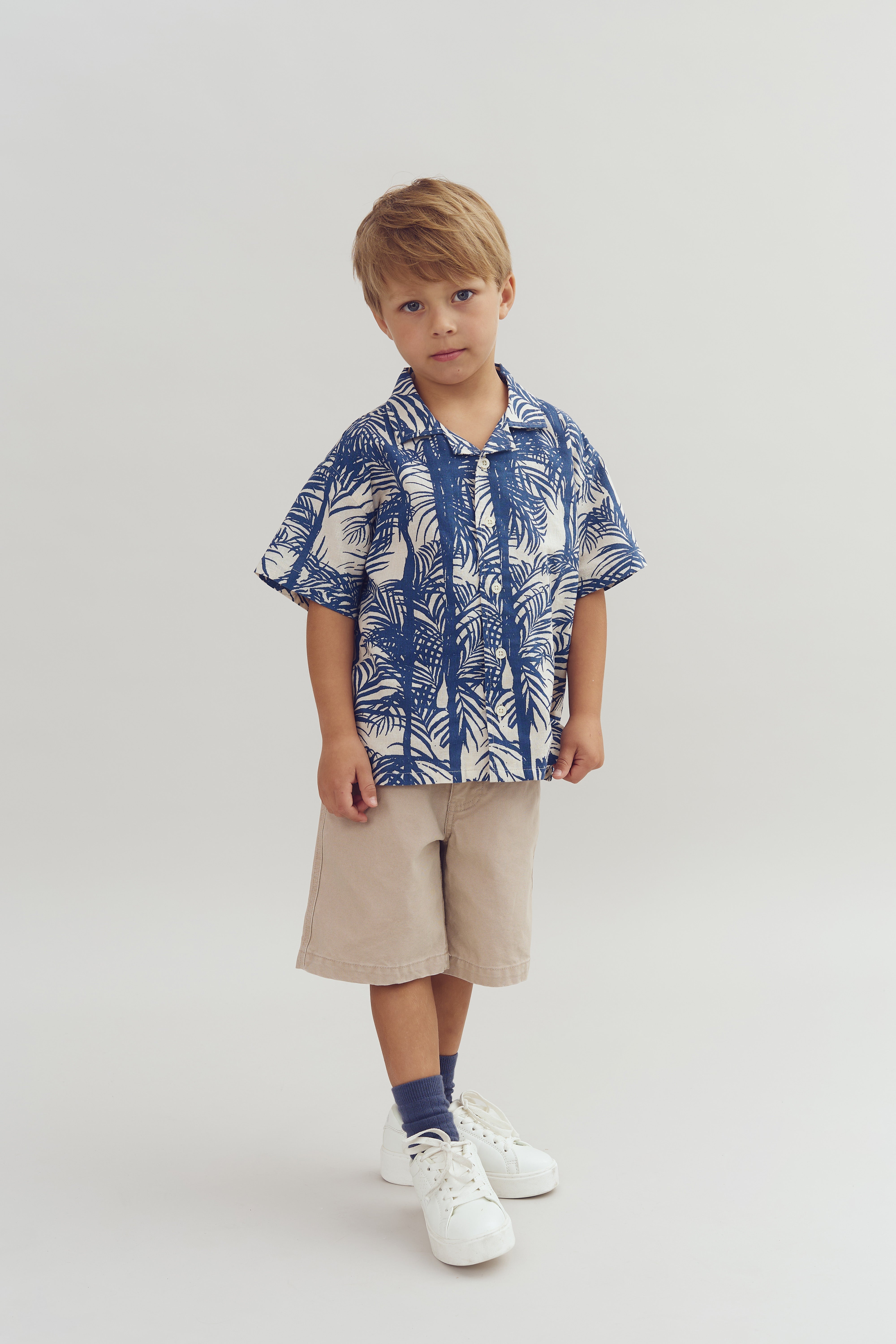 A young child stands wearing a blue patterned shirt, beige shorts, blue socks, and white sneakers. The background is a plain, light color.