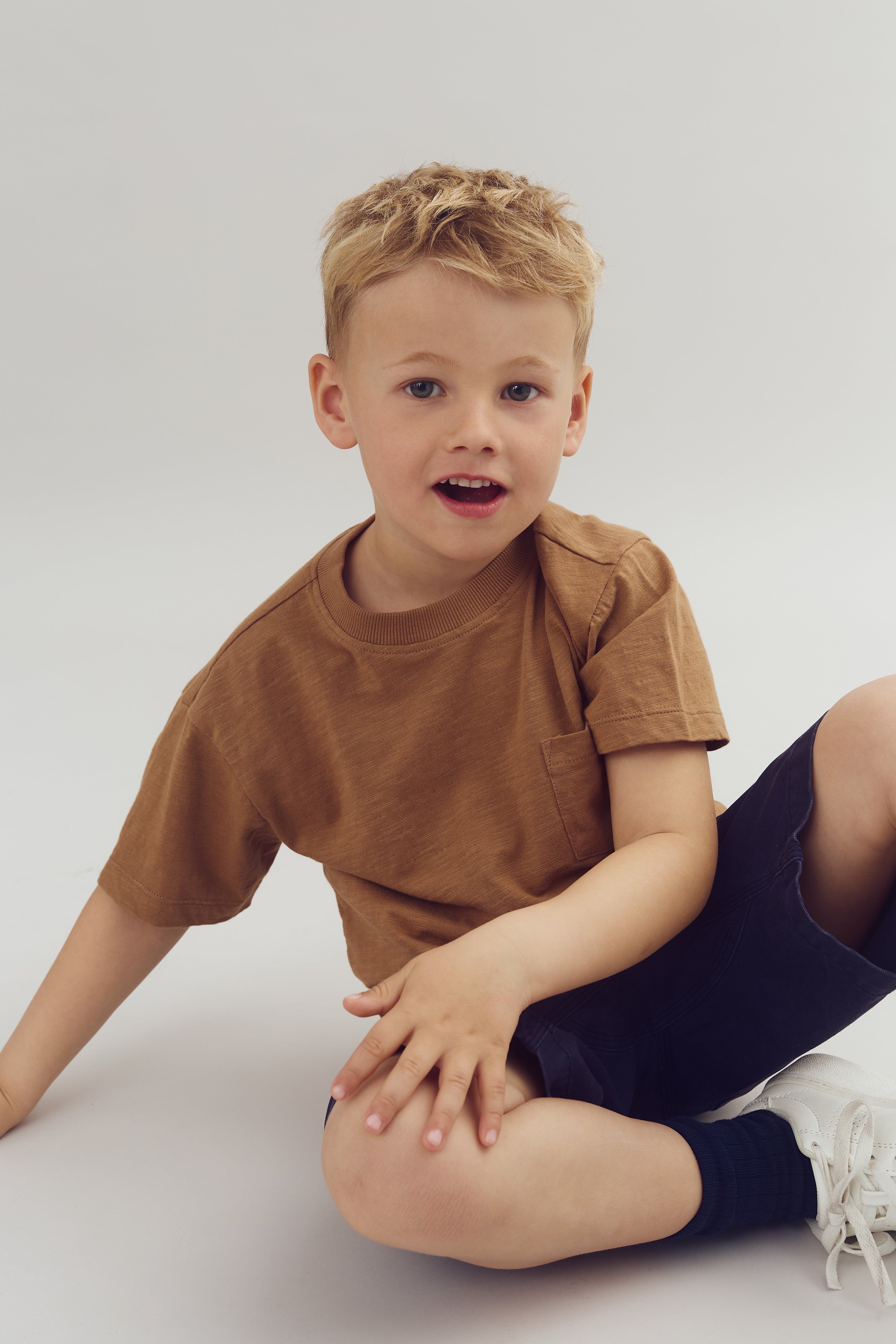 A young child sits cross-legged, wearing a brown T-shirt and dark shorts, smiling slightly. The background is a plain, light color, emphasizing a simple and relaxed setting.