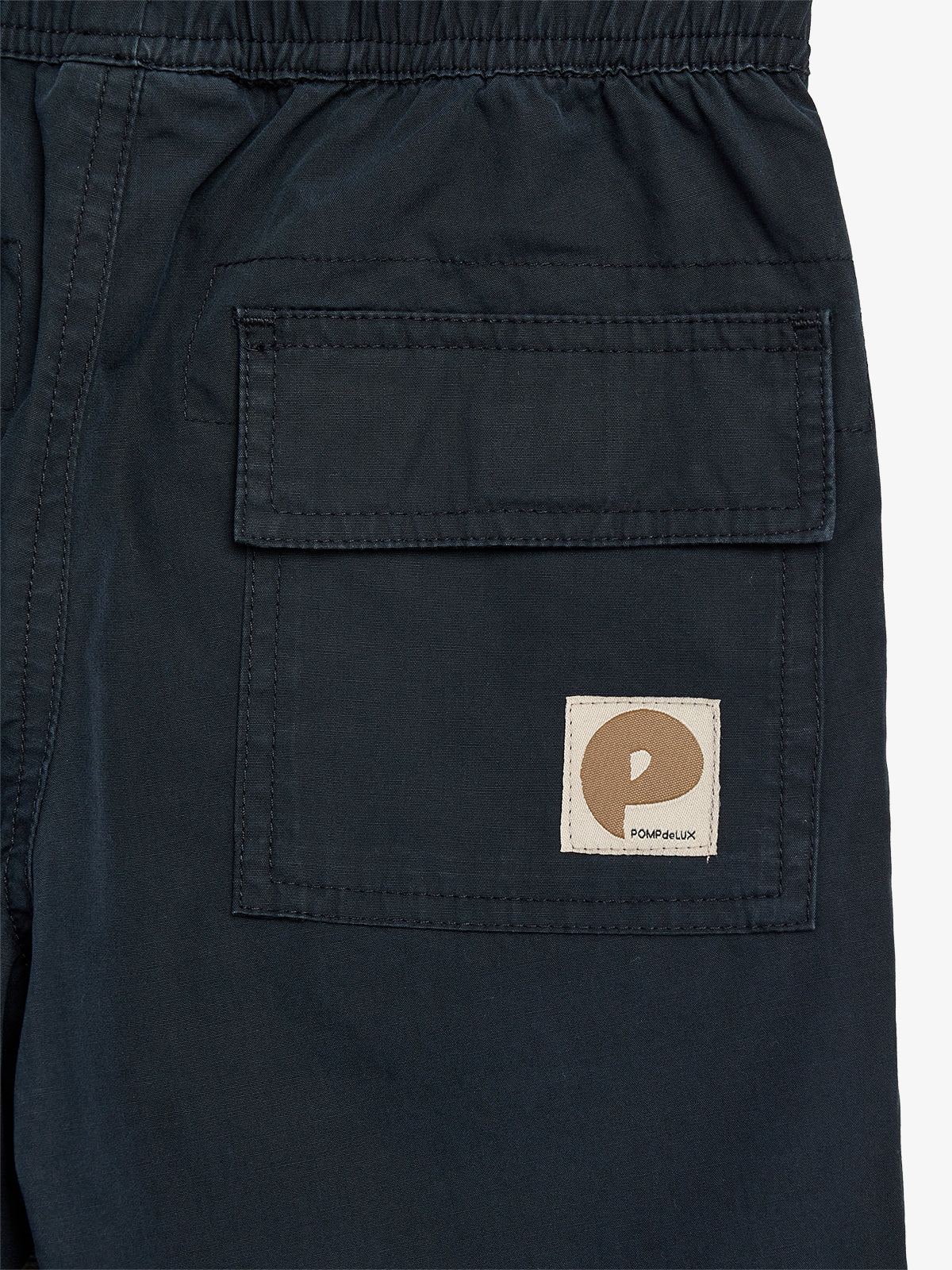 Dark fabric shorts feature a flap pocket with a light brown patch displaying a stylized "P." The text "POMPdeLUX" is beneath the patch, emphasizing branding.