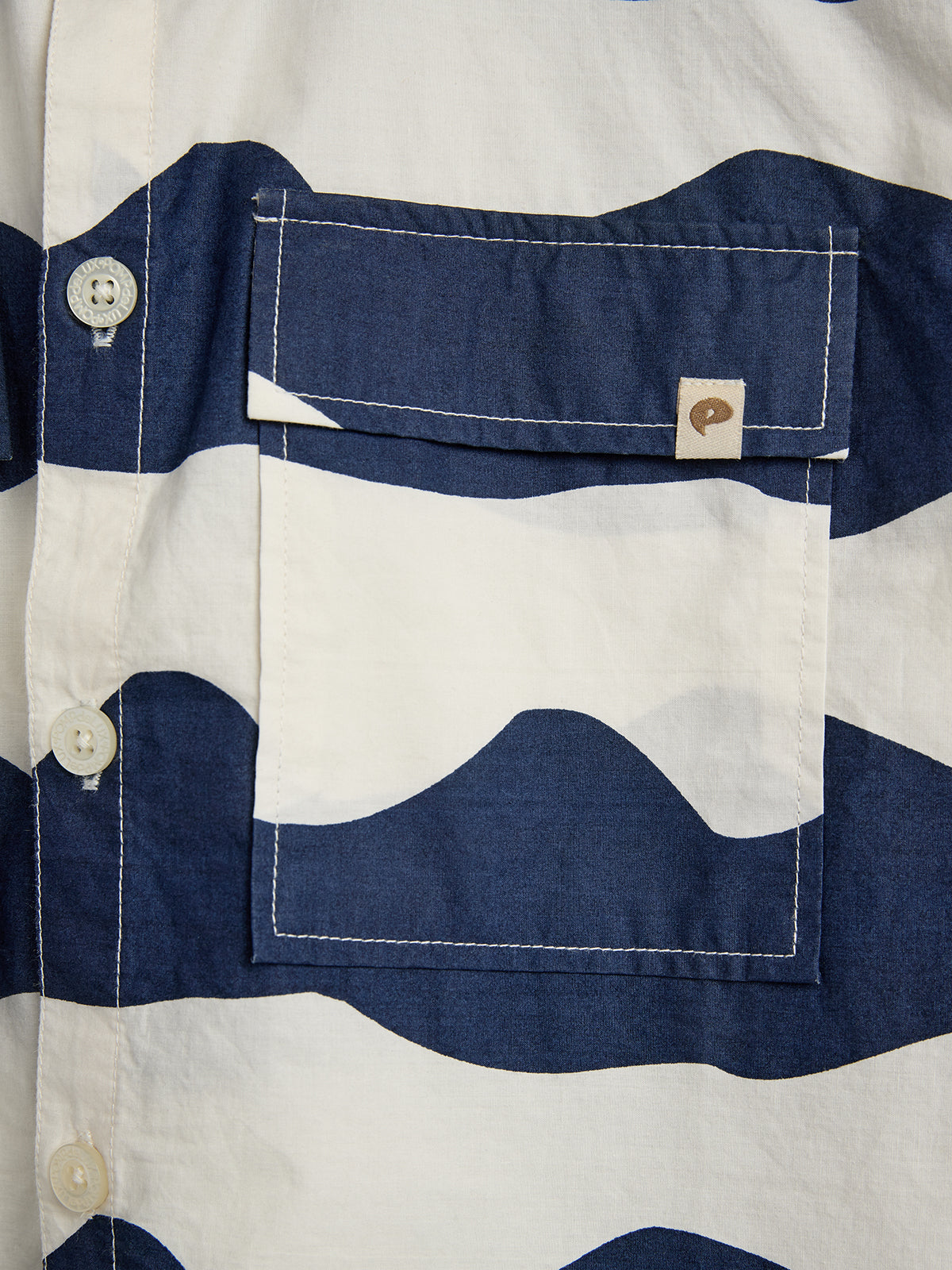 A shirt pocket with a flap in a wavy blue and white pattern. It has a button and a small tag with a stylized letter "p" in the top right.