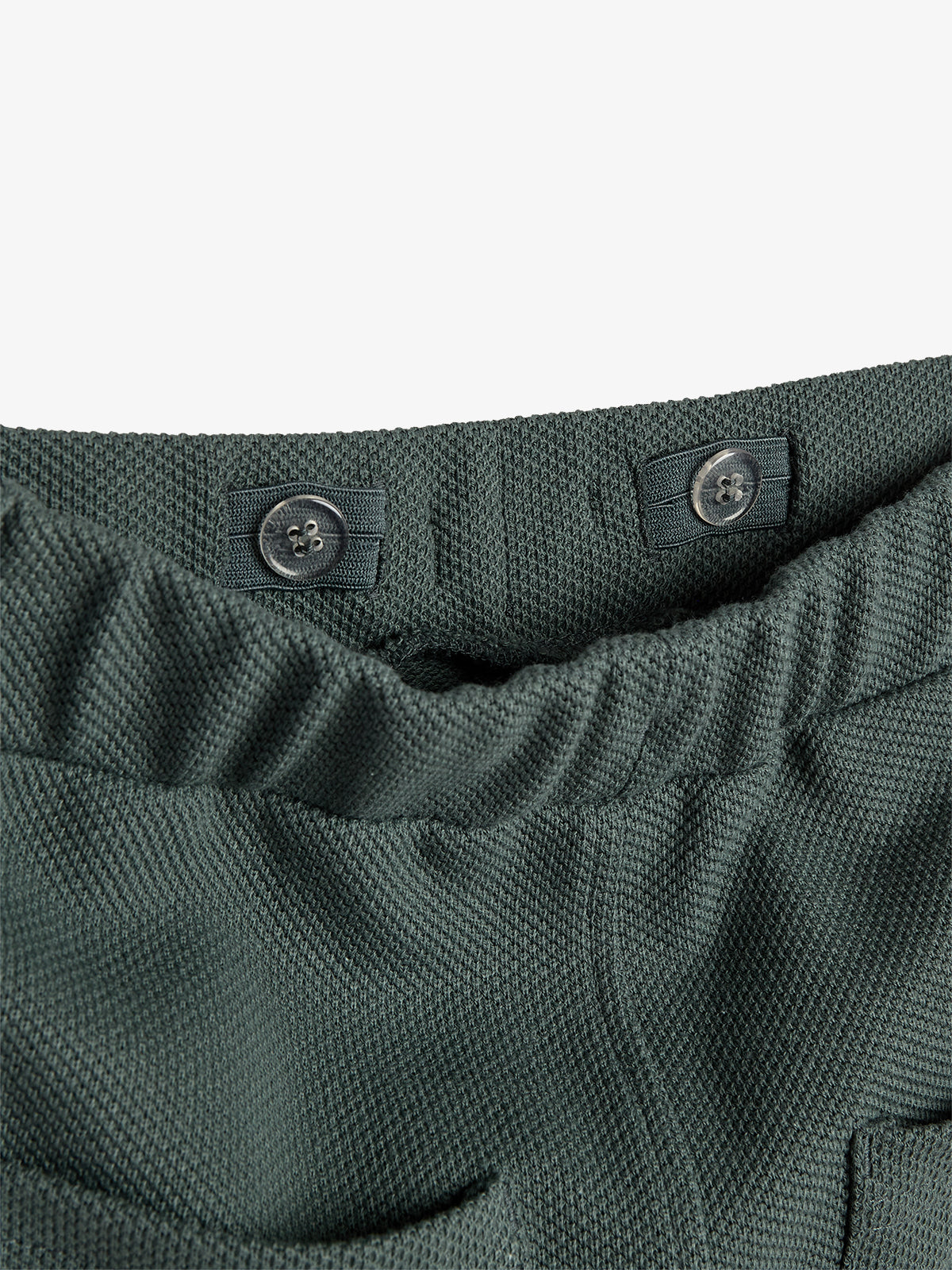 Dark green textured fabric waistband with two buttons, set against a plain background. The elastic waistband features subtle stitching, creating a gathered effect for a flexible fit.