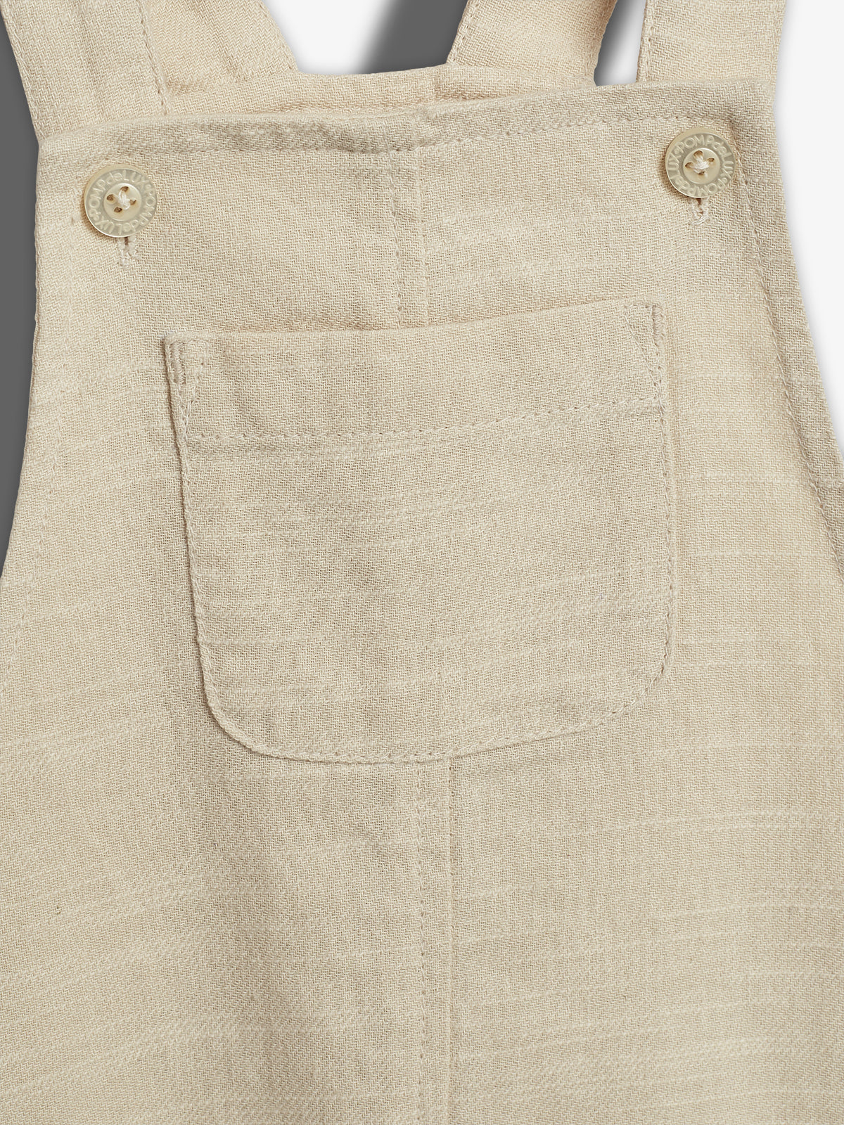 A beige fabric overall features a front pocket with parallel straps and two buttons. The buttons read "INSOUMUSE." The texture appears woven, providing a casual appearance.