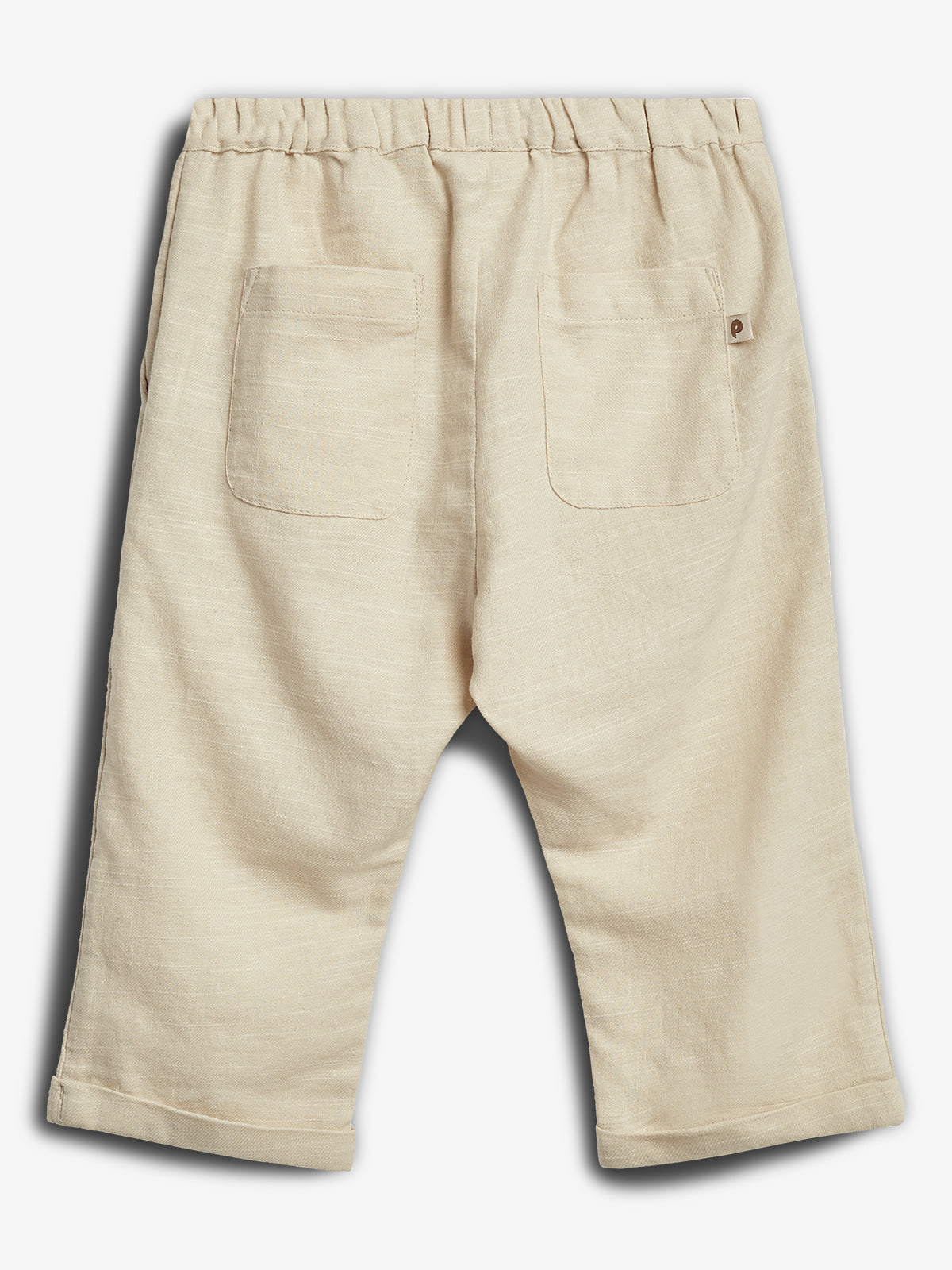 Beige linen shorts are laid flat, displaying two back pockets with stitching. The waistband is elasticized, suggesting comfort. A small label with a logo is attached near one pocket.