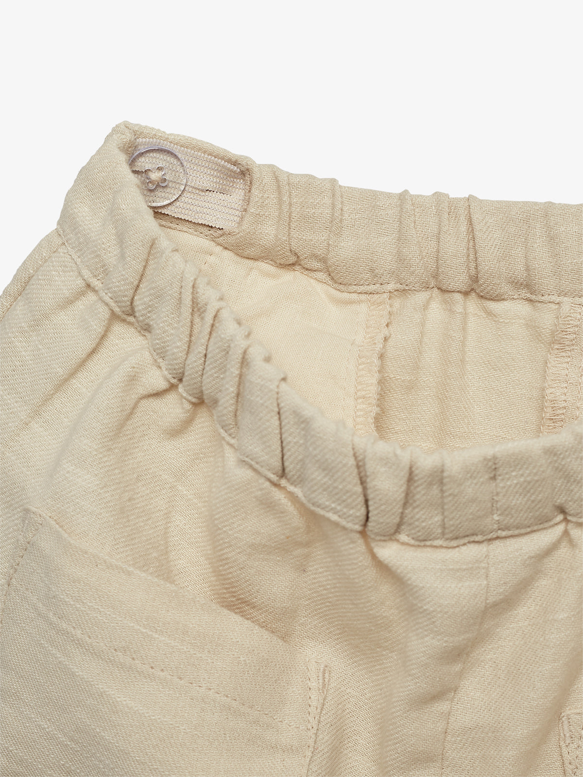 Beige pants feature an elastic waistband with a button, situated in a plain, close-up view. The fabric texture is visible, highlighting the pants' casual style.