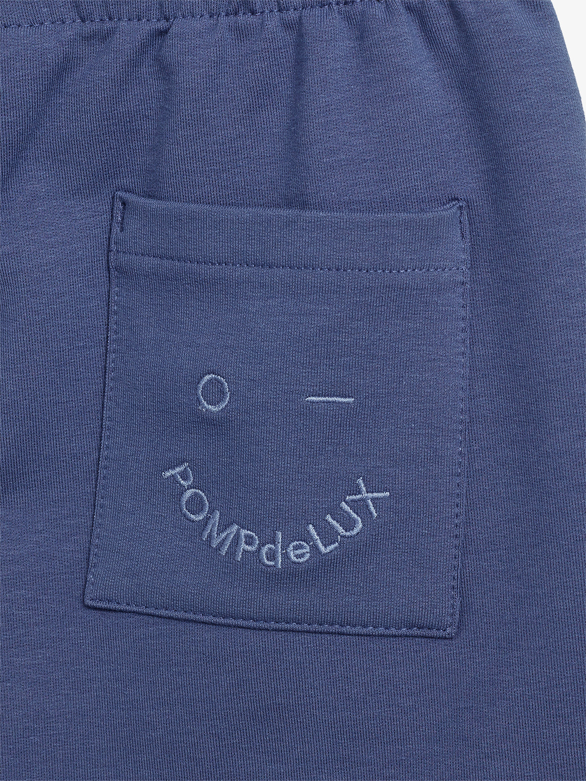 Blue fabric features a pocket with embroidery. The design includes a simple face and text: "POMPdeLUX". The fabric appears gathered at the top, suggesting an elastic waistband.