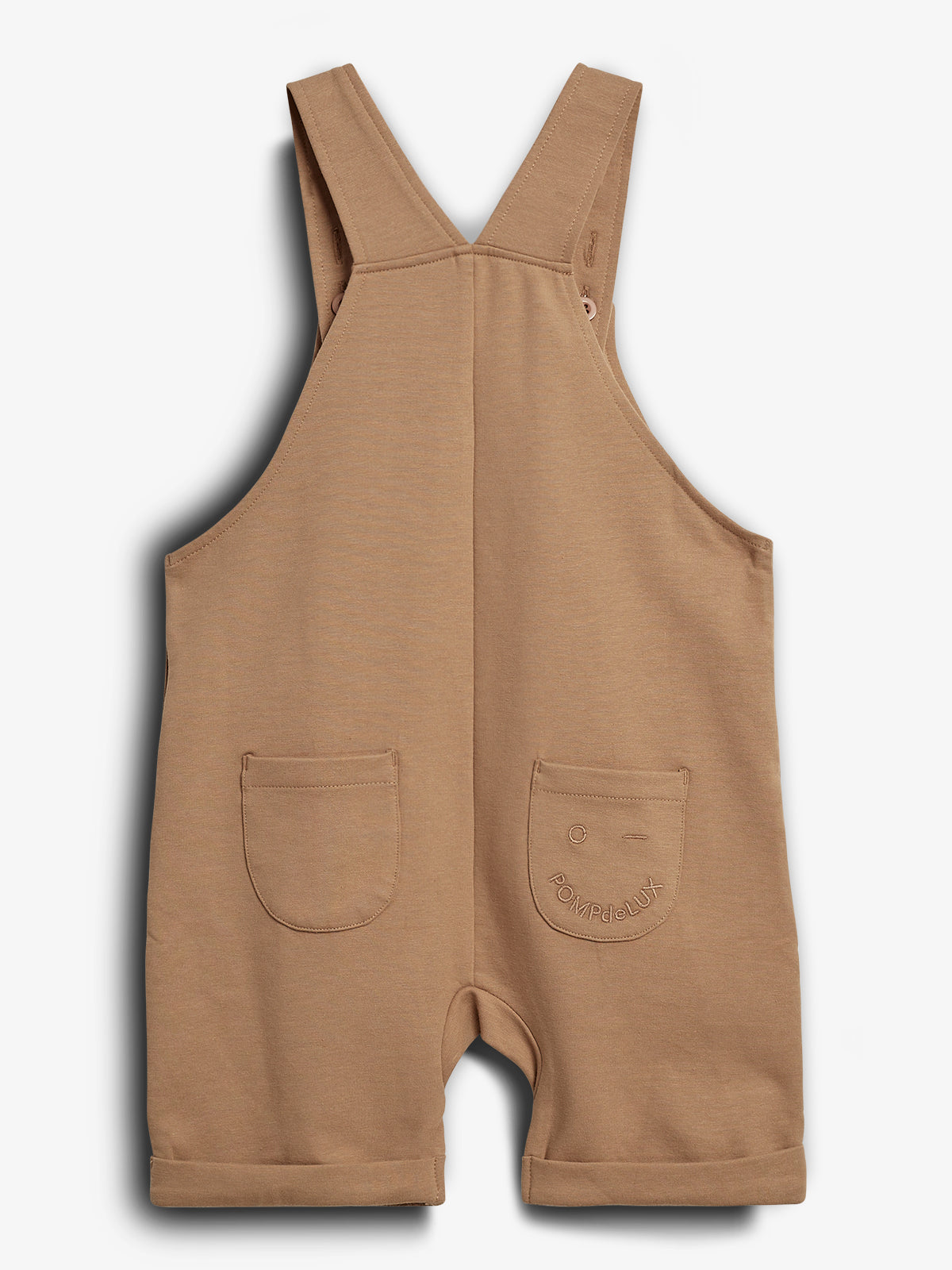 A tan-colored toddler romper with two front pockets, featuring an embroidered face and the text "POMPdeLUX" on the right pocket. It has wide straps and is displayed on a plain background.