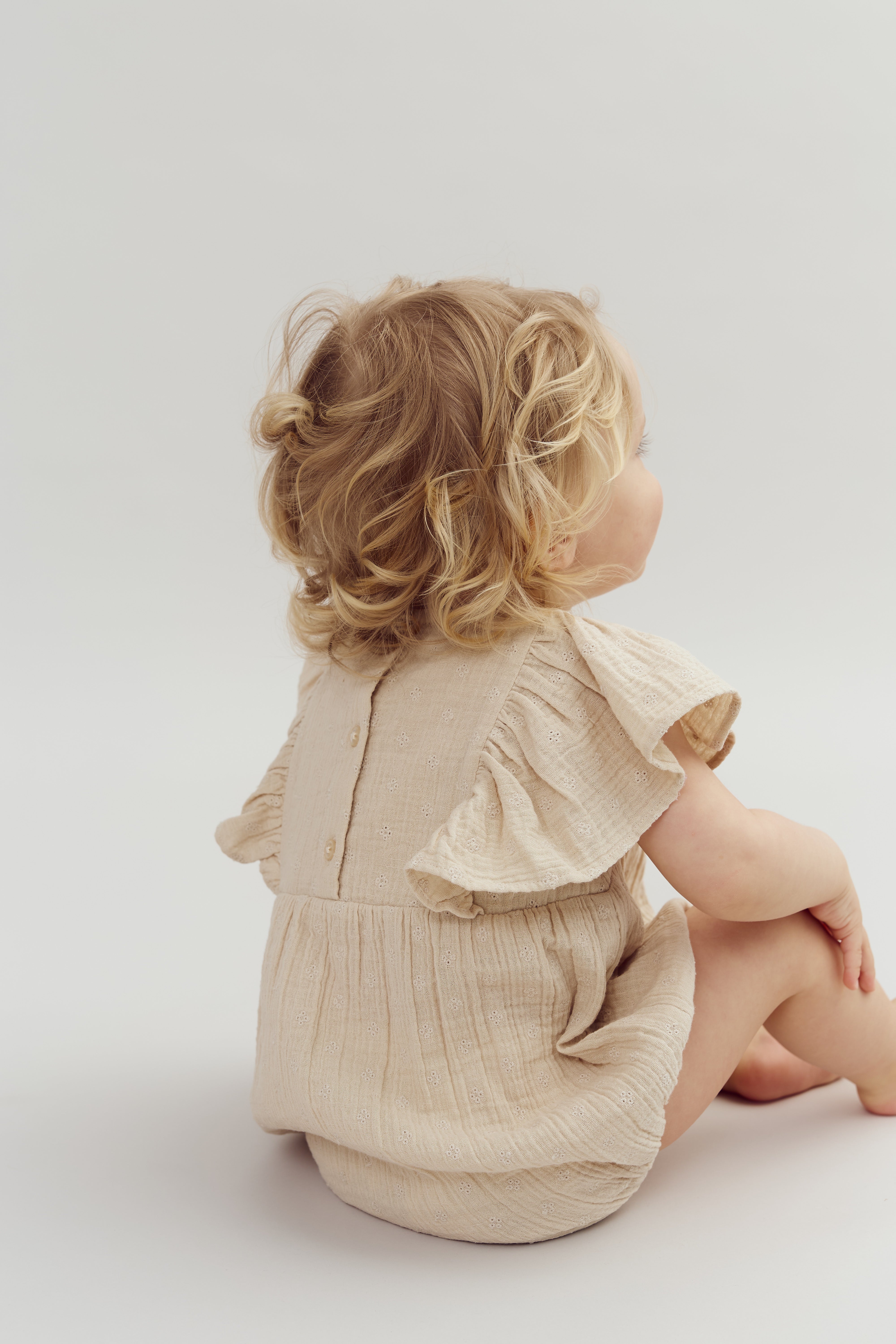 A toddler with curly blonde hair sits on the floor, wearing a beige outfit with ruffled sleeves, in a bright, minimalistic setting.
