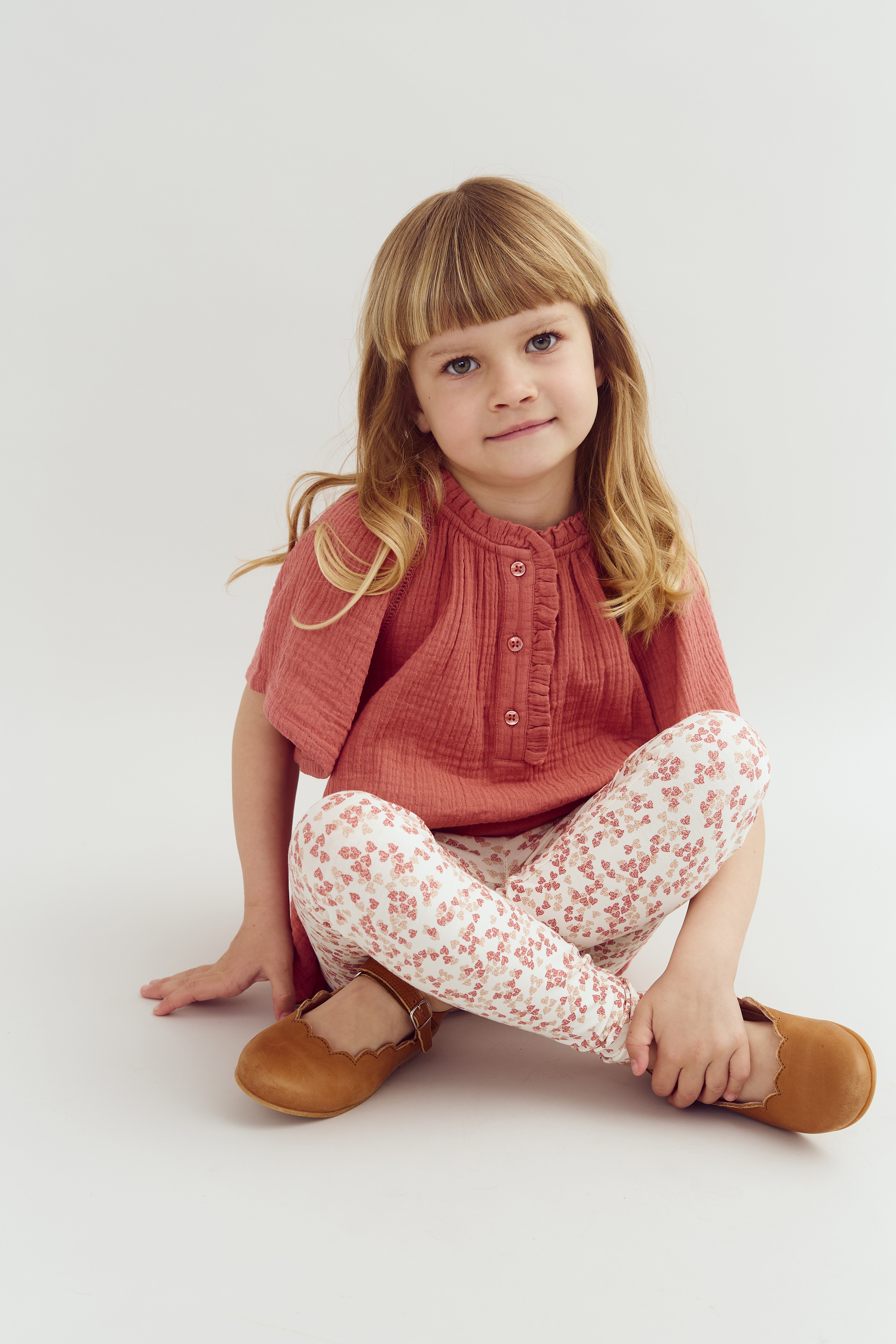 A young girl is sitting on the floor, wearing a coral top and floral-patterned leggings. Her legs are crossed, and she is smiling slightly against a plain white background.