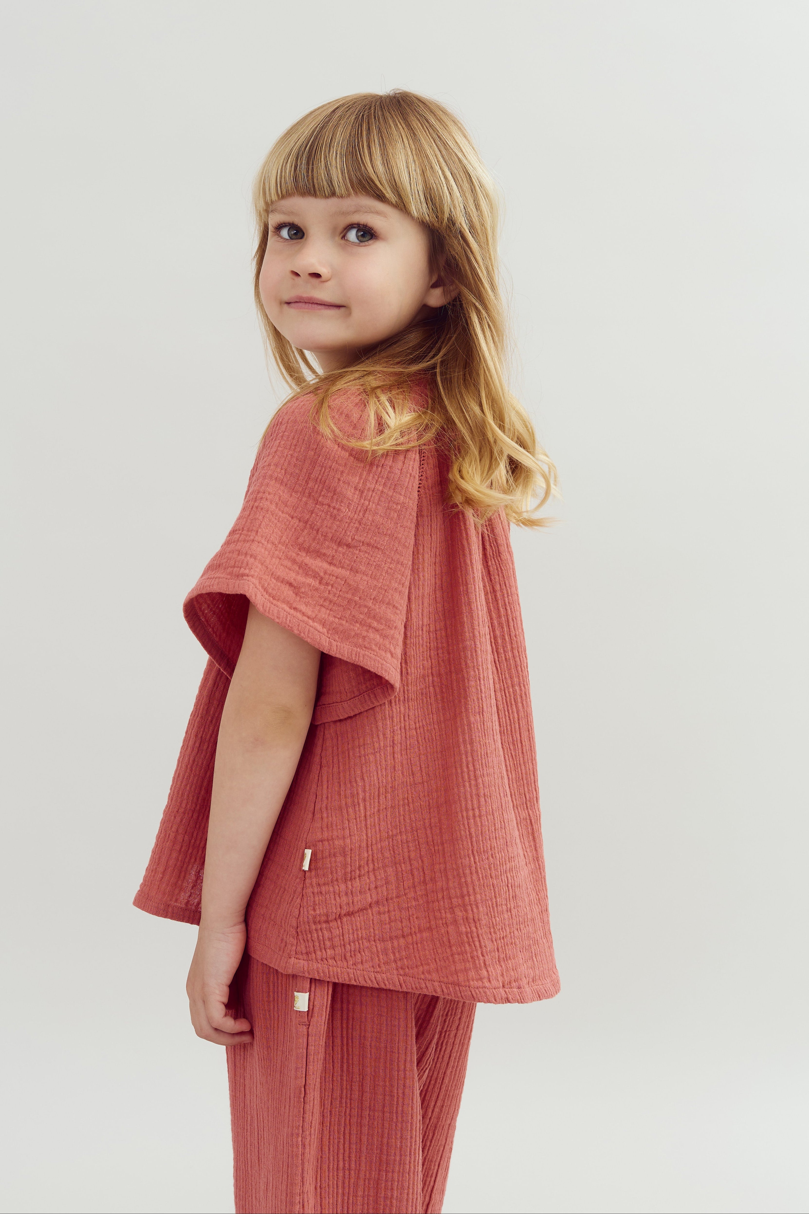 Child wearing a textured, coral-colored outfit looks over their shoulder, standing in front of a light gray background.