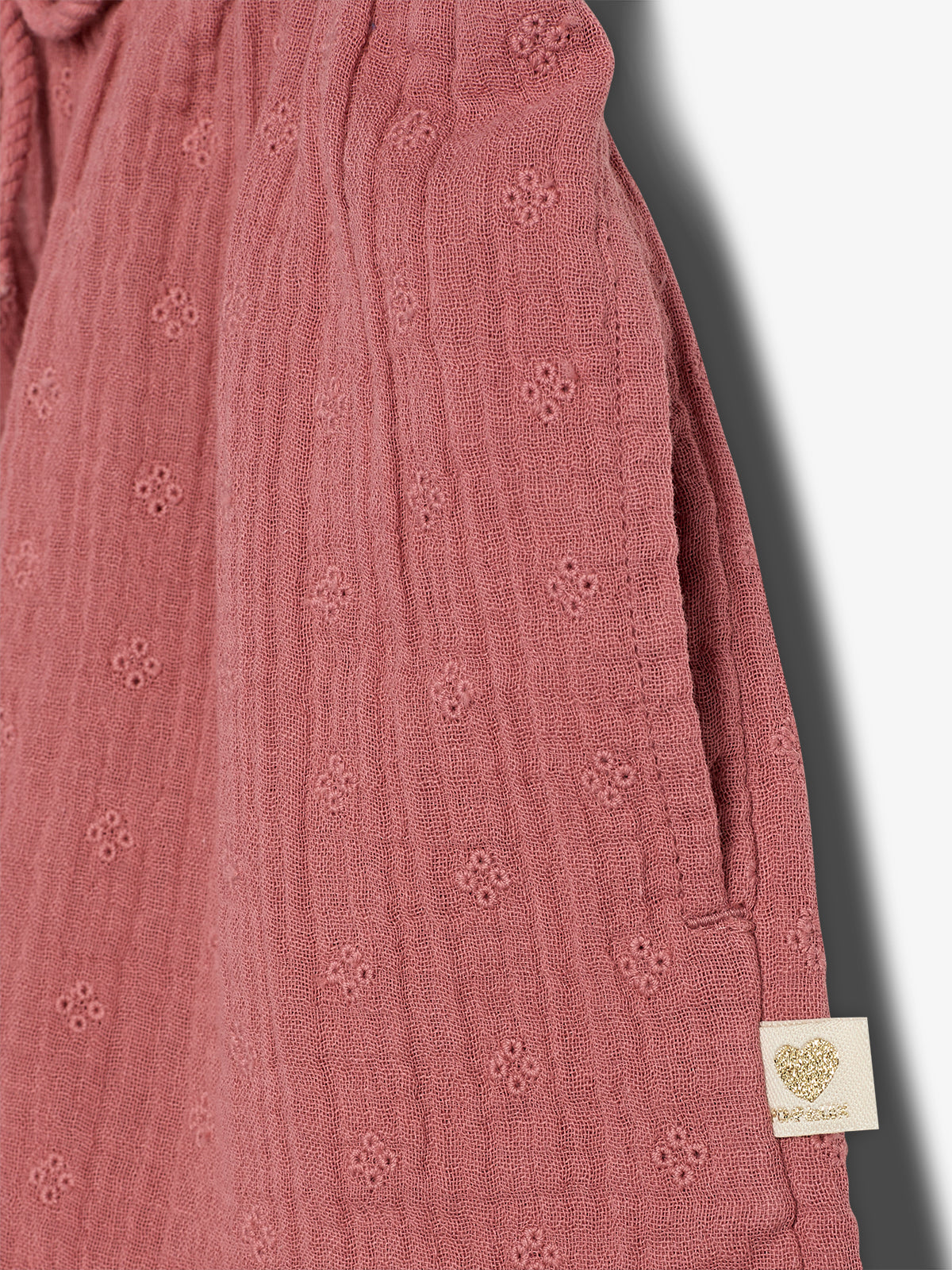 Pink textured fabric with small floral patterns features a cream tag displaying a gold heart. The fabric is draped vertically, casting a shadow on a white background.