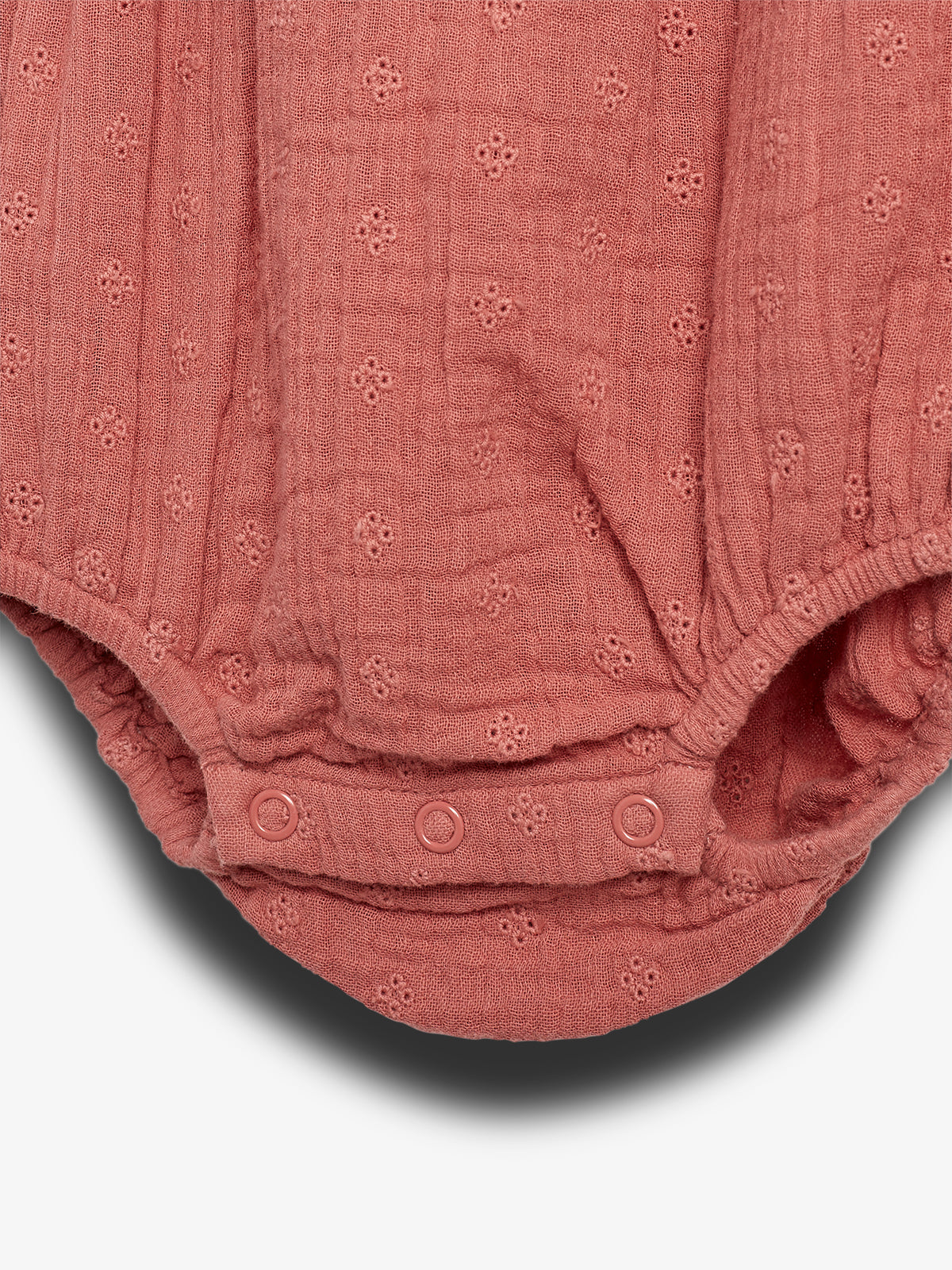 A textured, pink baby romper features small eyelet patterns and snap closures at the bottom, lying flat against a plain, light background.