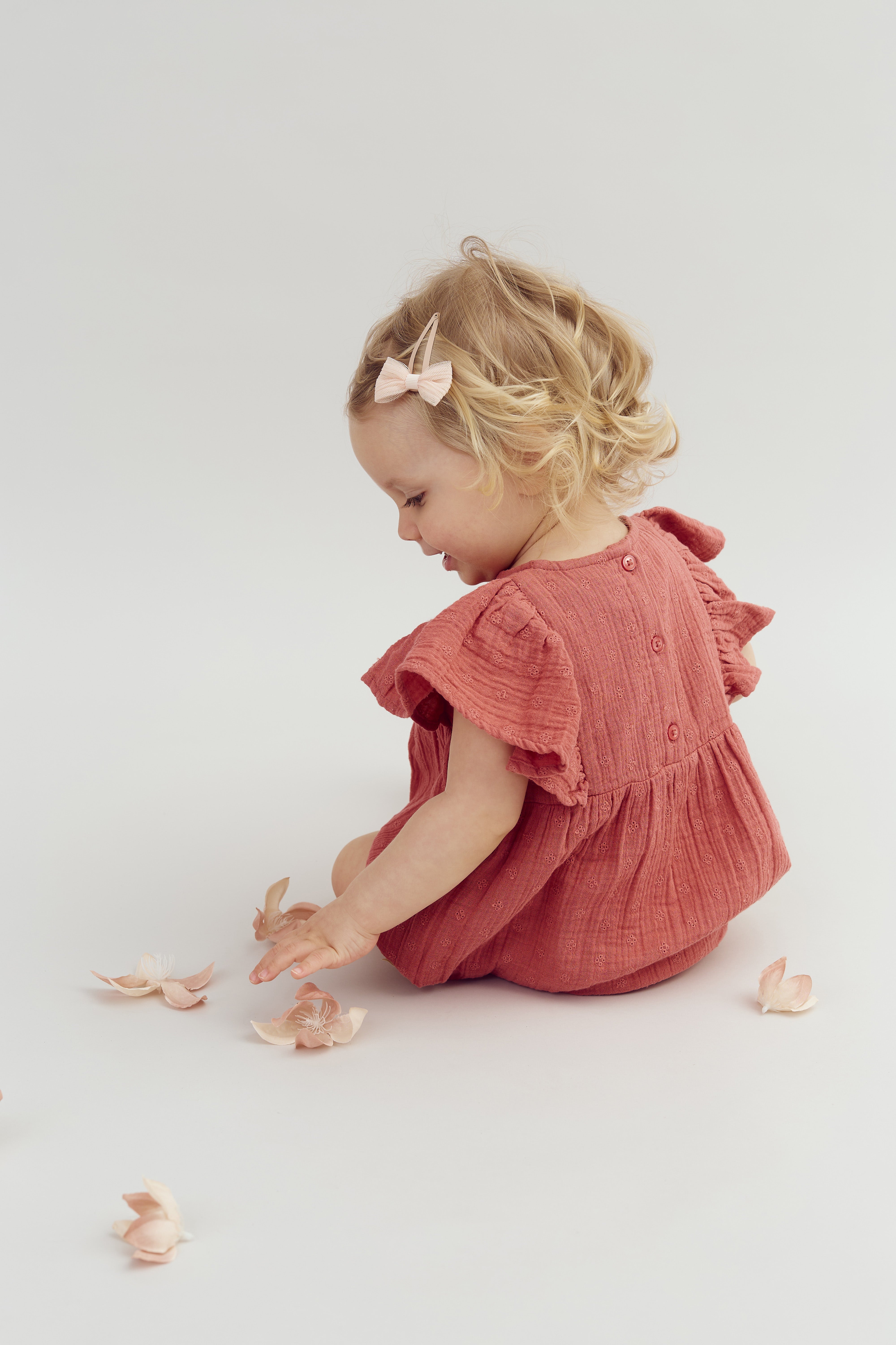 A toddler in a pink dress and hair bow is sitting and reaching for scattered pale pink flower petals on a plain, light background.
