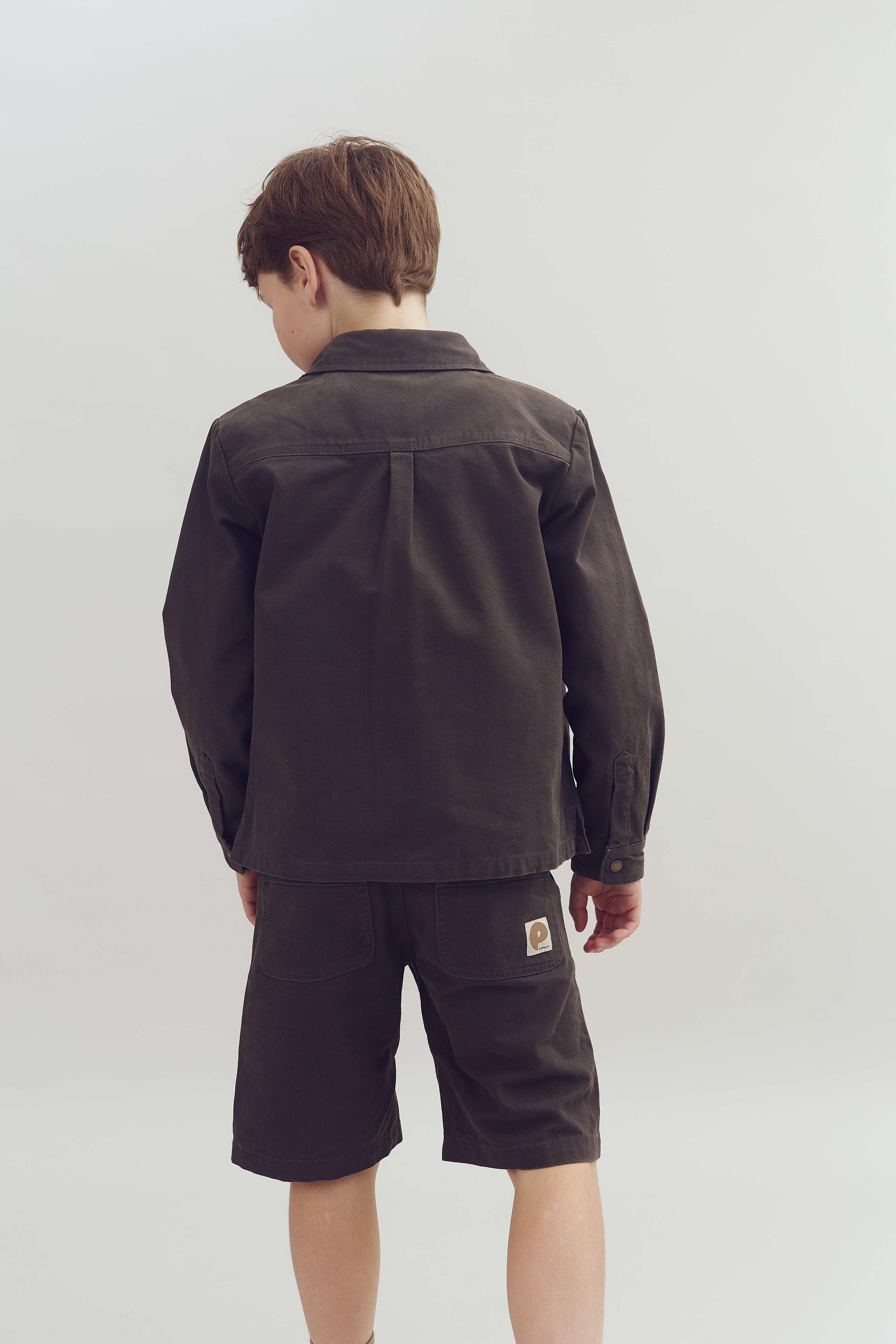 A person in dark brown jacket and shorts stands facing away in a neutral, light-colored setting. The shorts have a small "e" label on the back pocket.