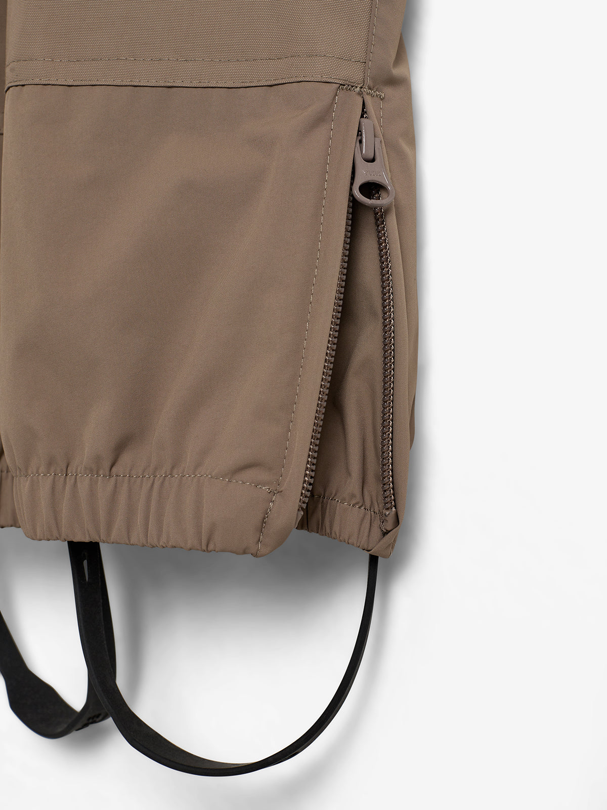 A tan jacket at the hem features a partially opened vertical zipper, revealing black strap loops below, against a plain white background.