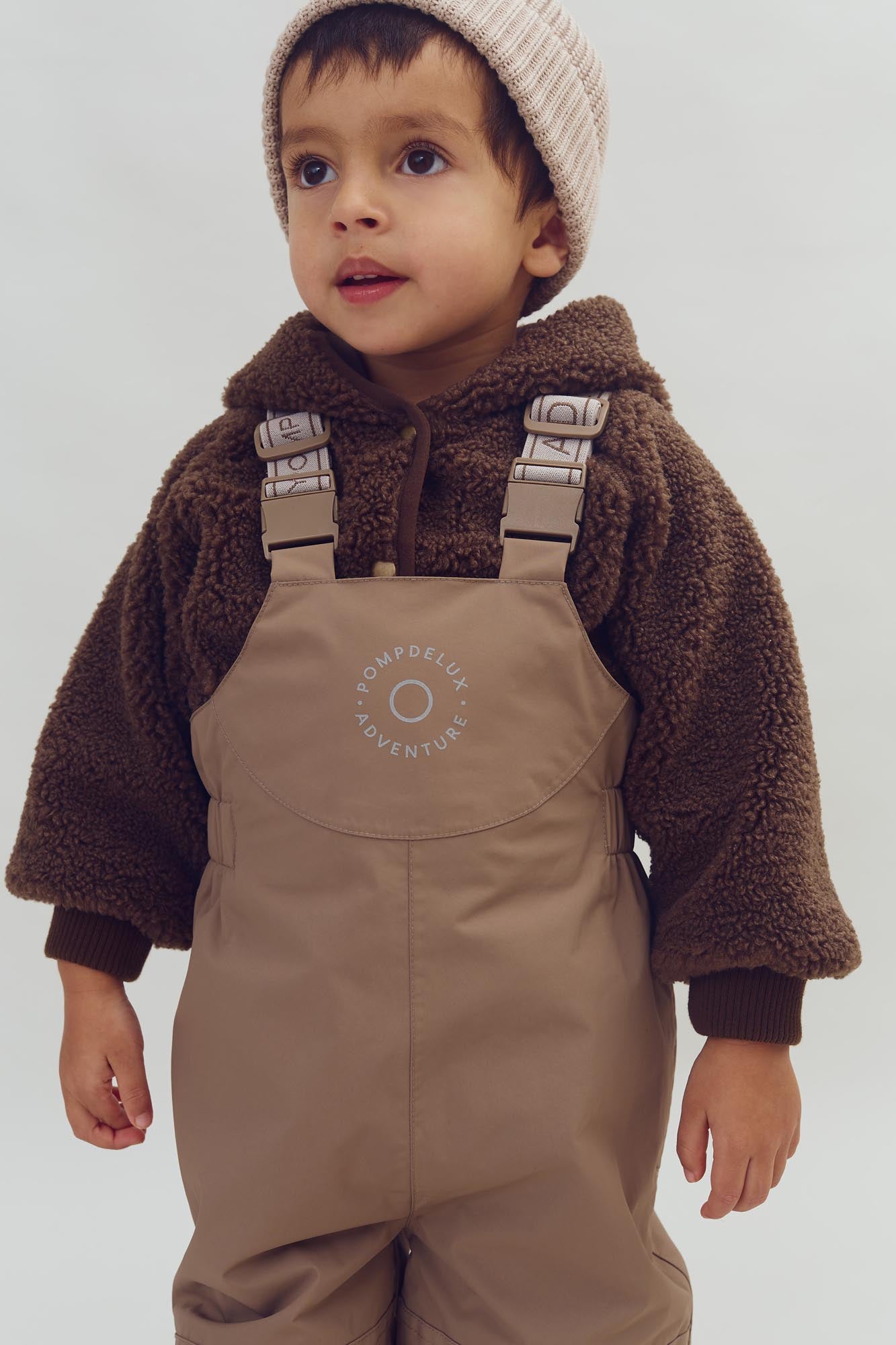 Child stands wearing a brown fleece jacket, beige overalls, and a knit beanie. The overalls feature the text "POMPDELUX ADVENTURE" in a simple indoor setting.