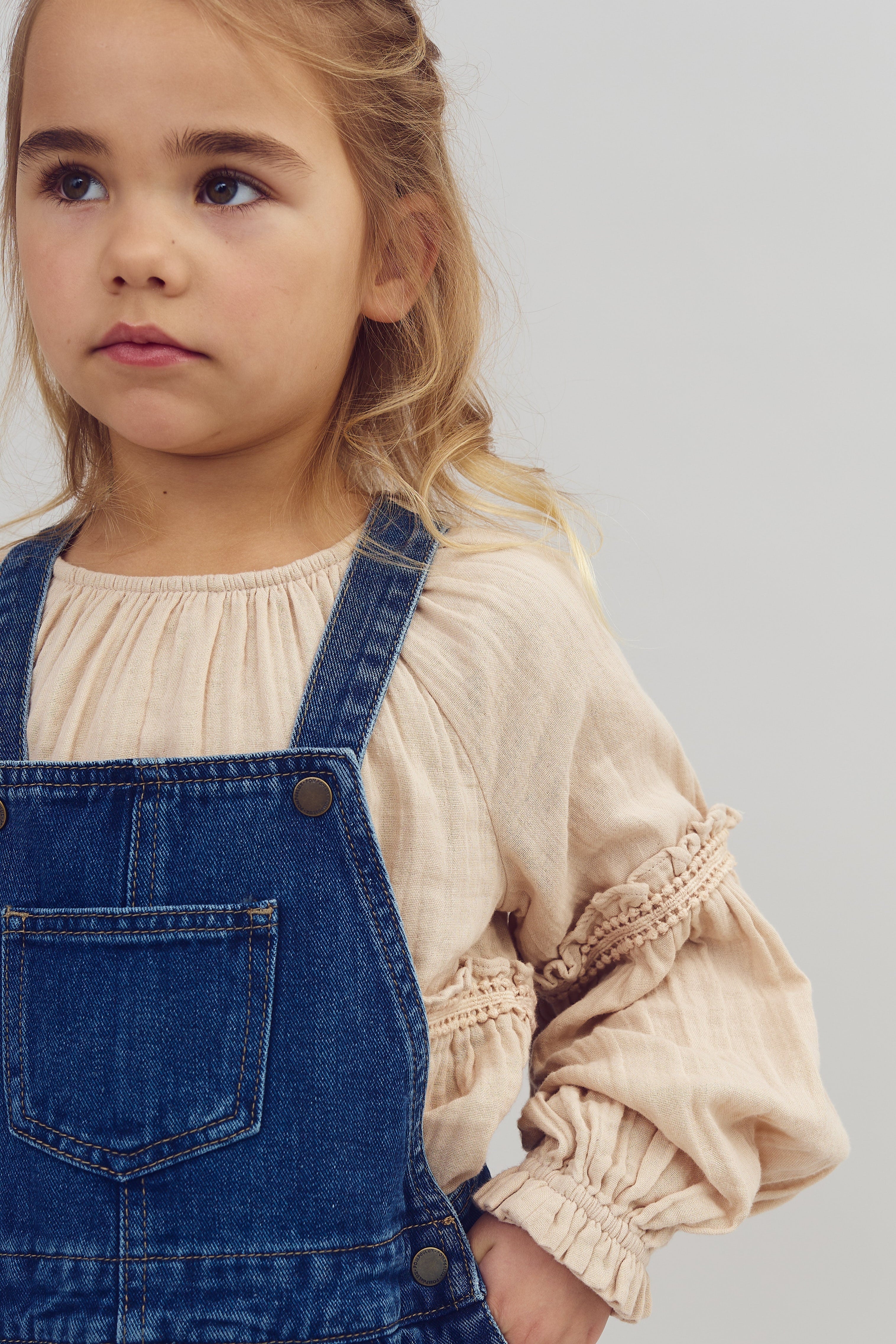 A young child stands in a blue denim overall and beige blouse, looking slightly to the left against a plain, light background.