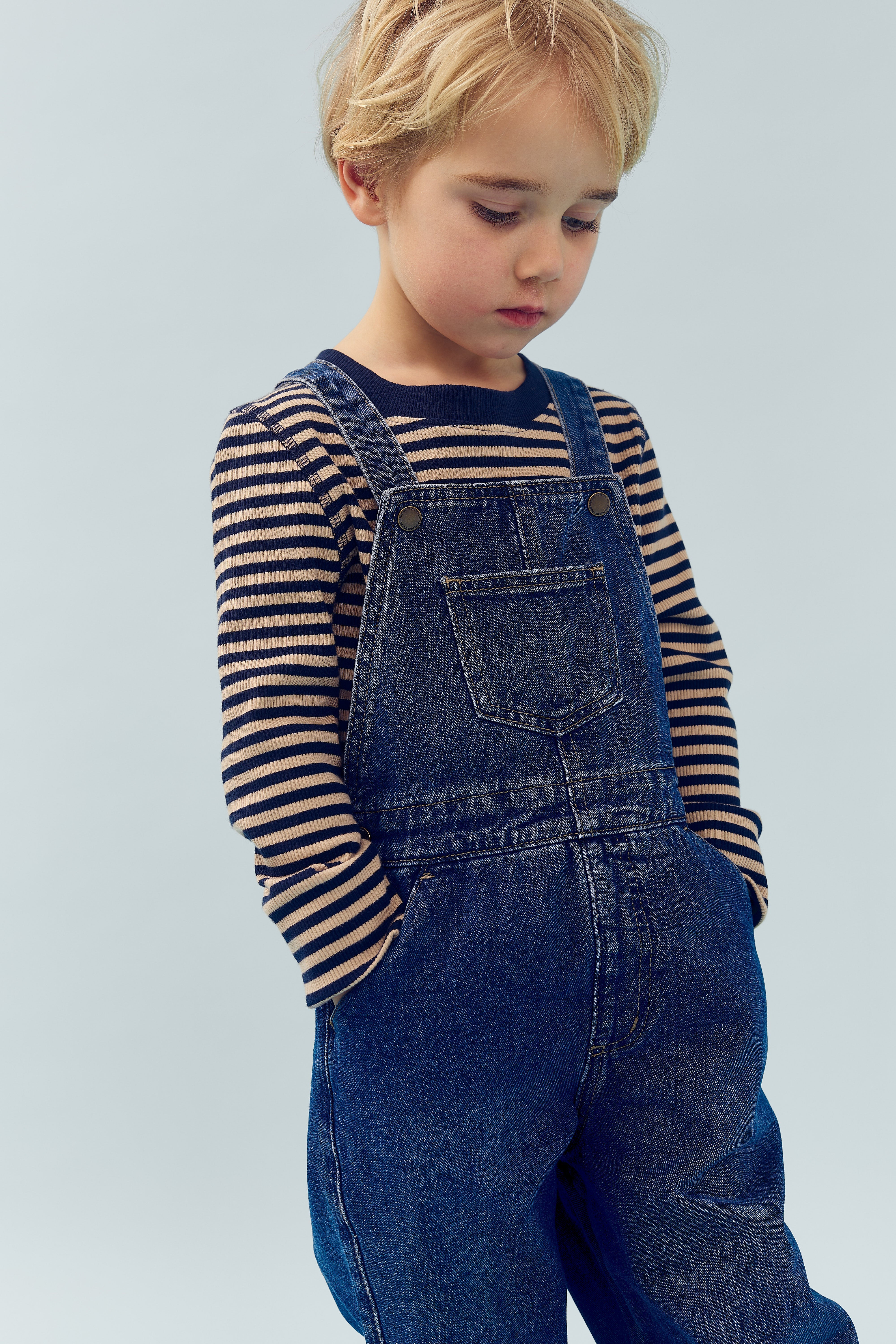Child with blond hair stands wearing denim overalls and a striped shirt, hands in pockets, against a plain light background, appearing thoughtful or contemplative.