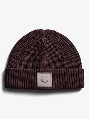 A knitted maroon beanie with a folded rim features a beige patch labeled "POMPdeLUX" and a simple face design, resting on a plain white background.