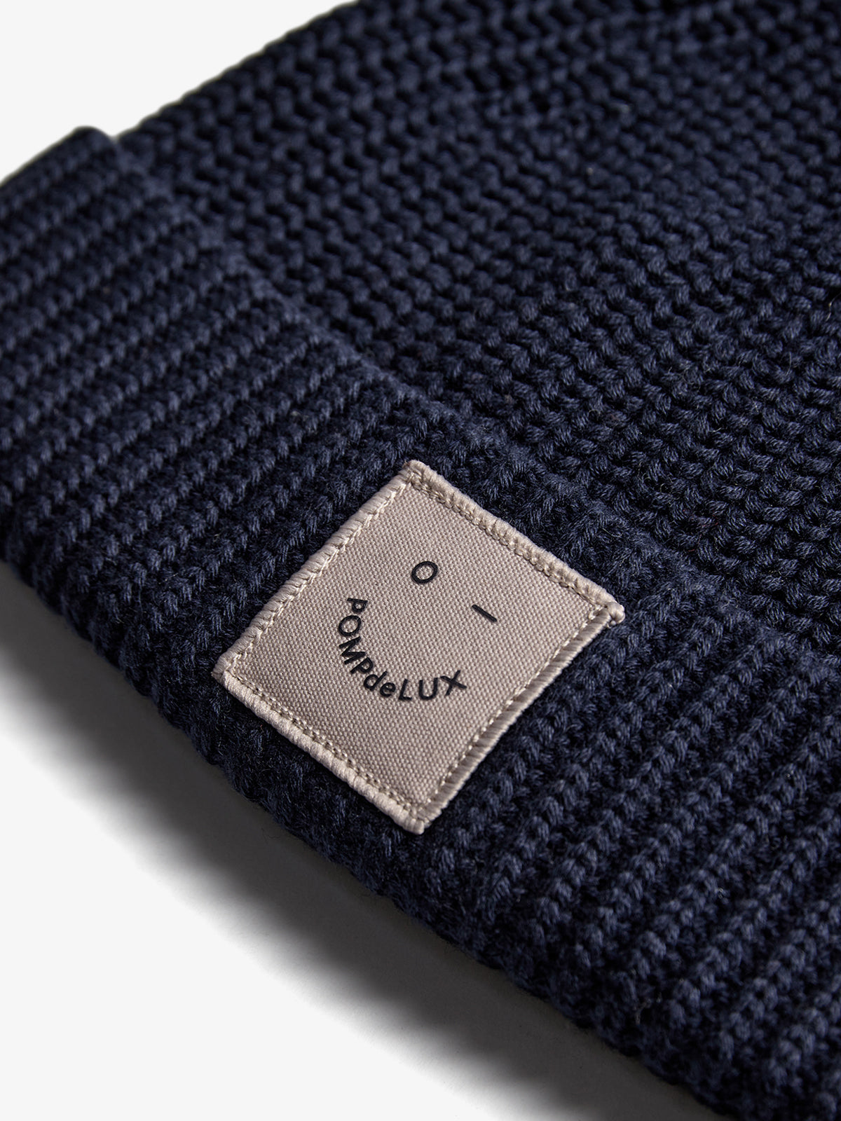 A navy knit beanie features a small beige patch displaying "POMPdeLUX" and a minimal smiley face design, resting on a neutral background.