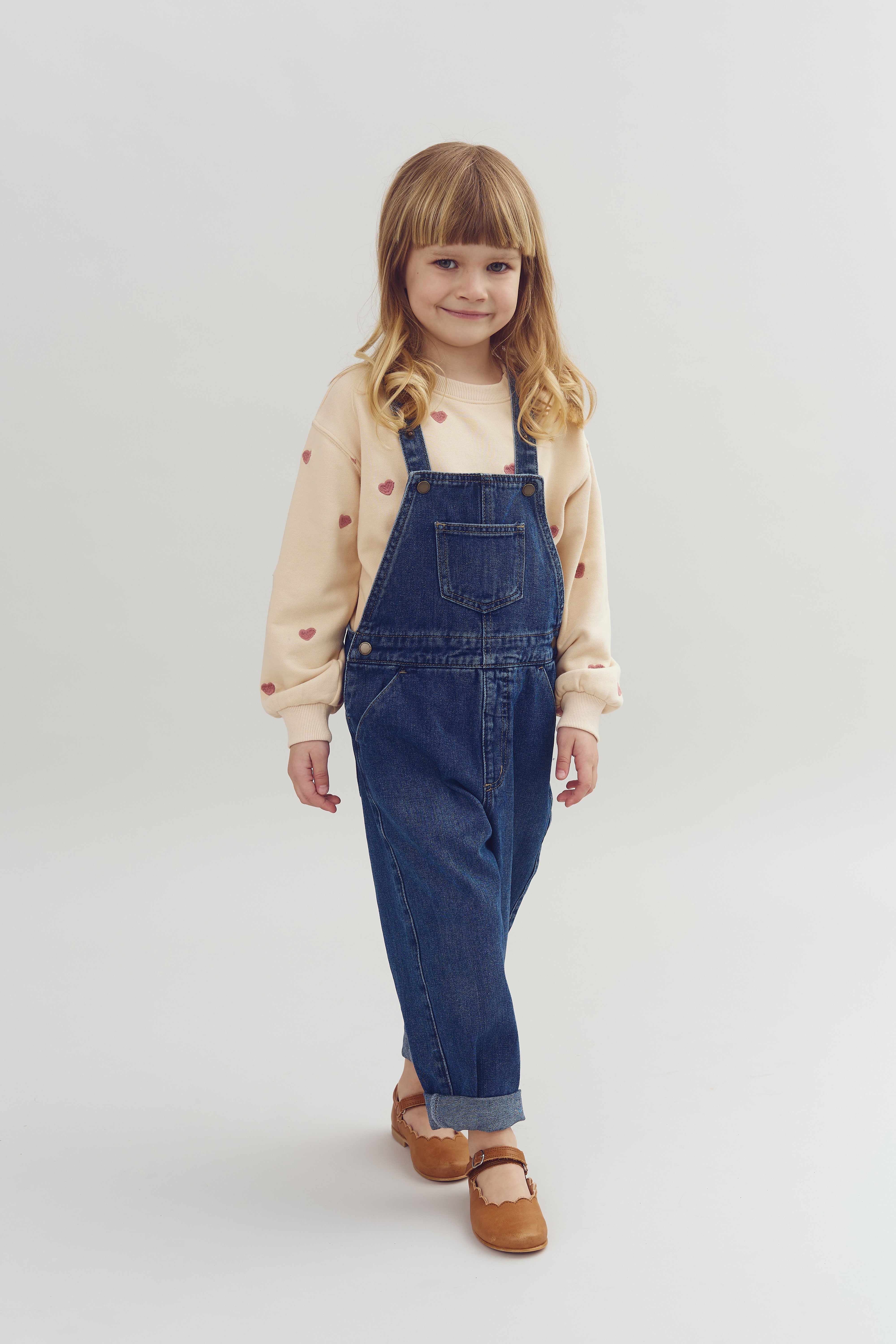A child wearing a light sweater with heart patterns and denim overalls stands smiling. They are in a neutral-colored studio environment, wearing brown shoes.