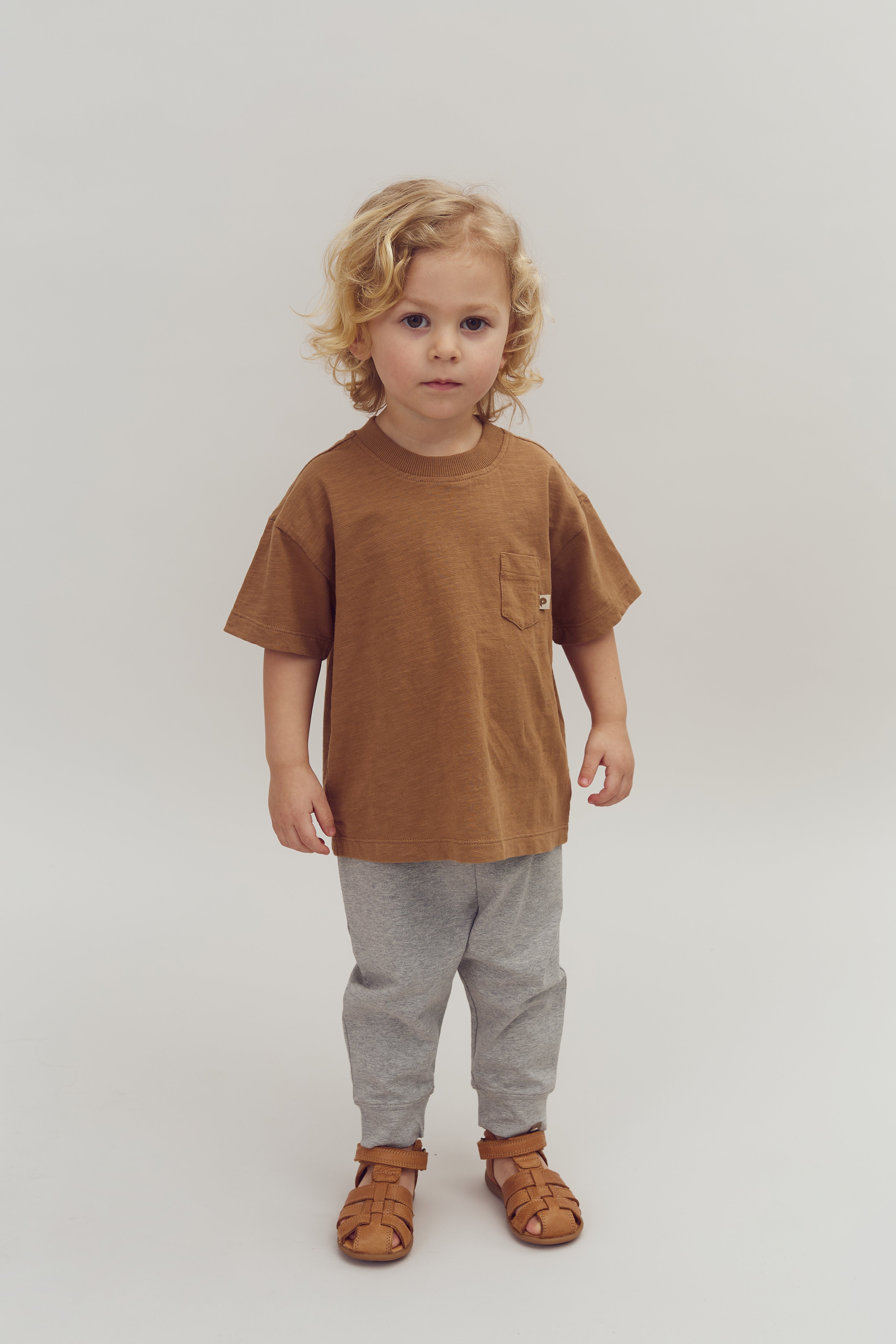 A child with curly hair stands still, wearing a brown shirt, gray pants, and brown sandals, set against a plain white background.
