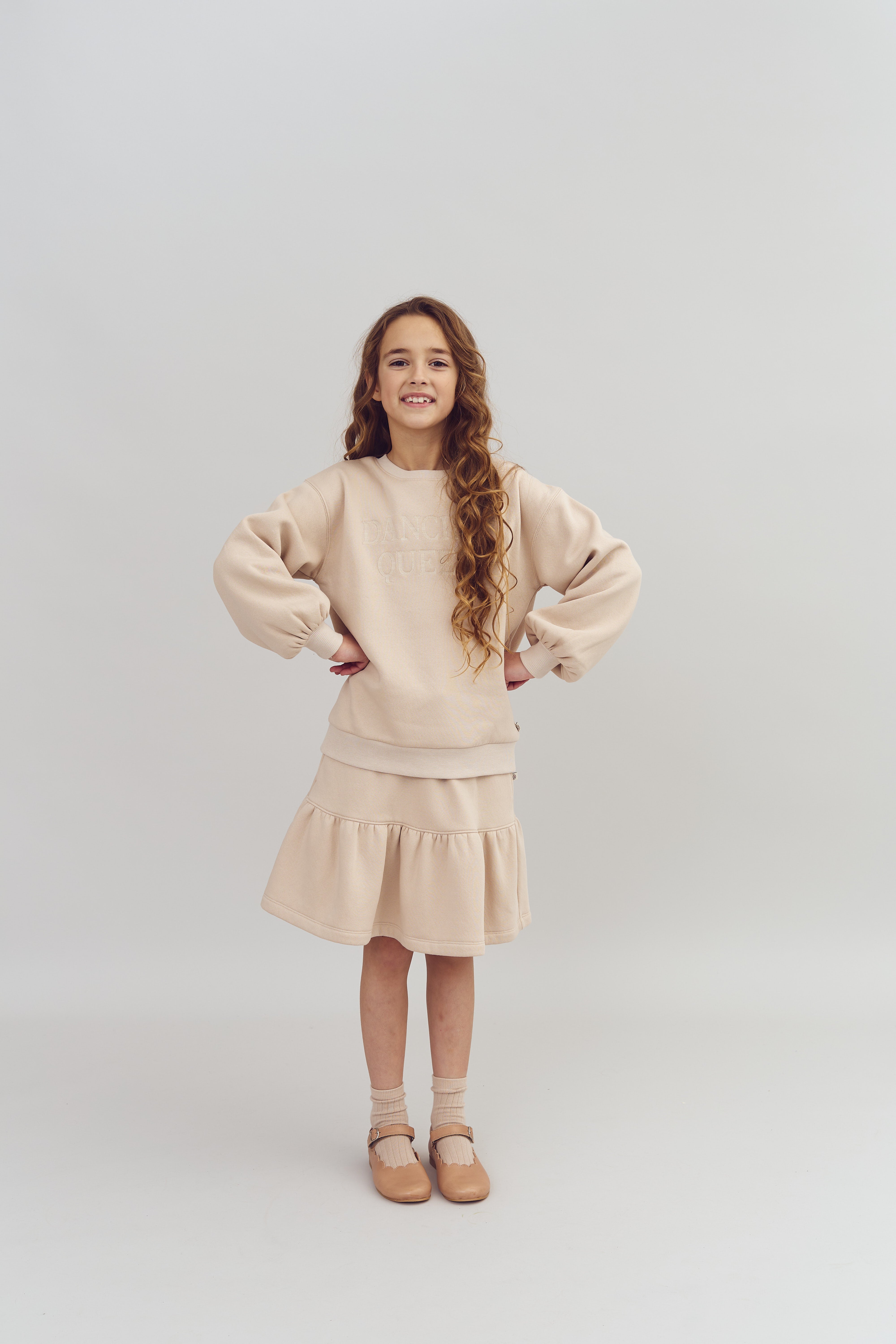 A young girl stands confidently with hands on hips, wearing a beige outfit reading "DANCE QUEEN," in a plain studio setting.