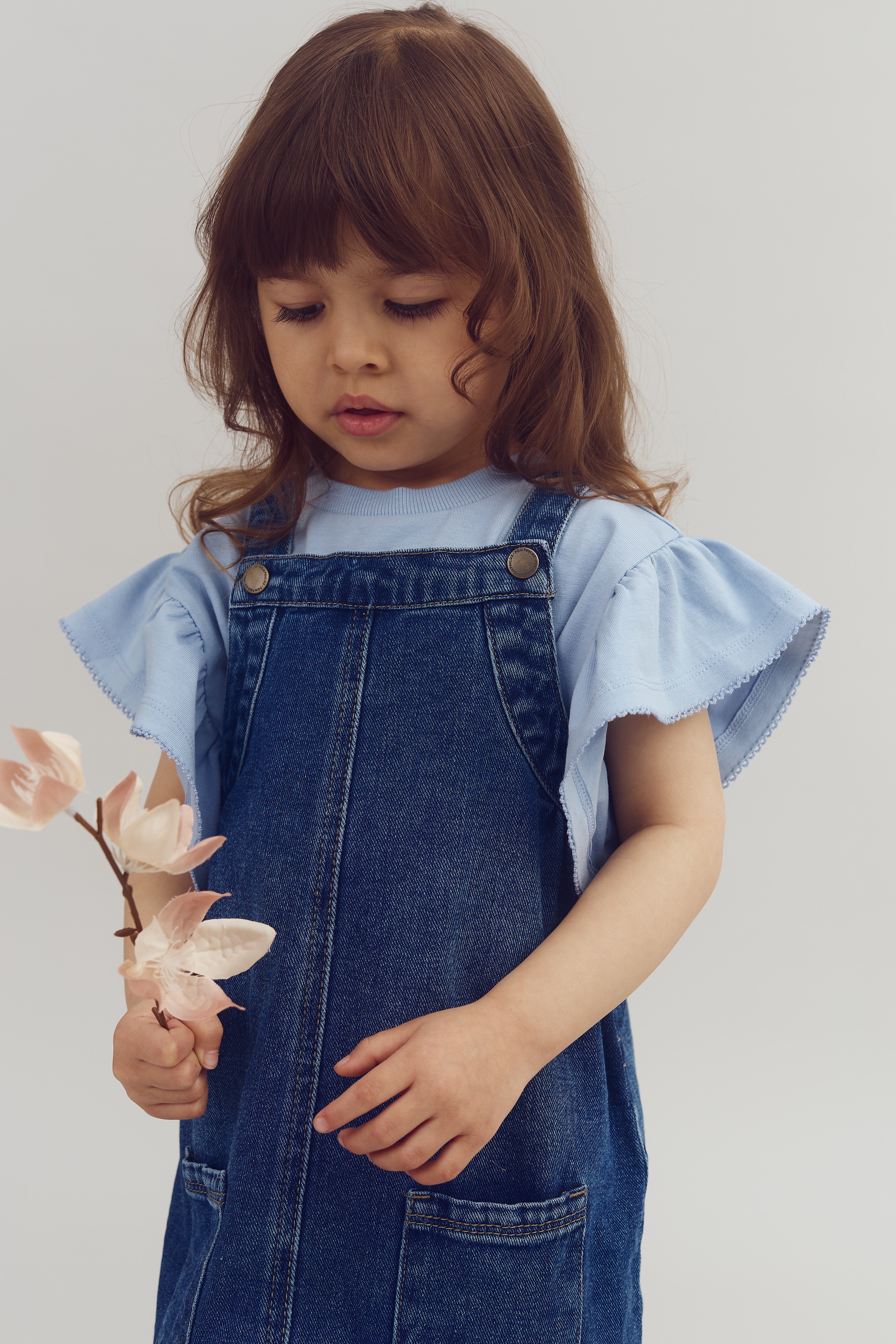 A young child holds a branch with pink flowers, wearing a denim dress over a light blue shirt, set against a plain off-white background.