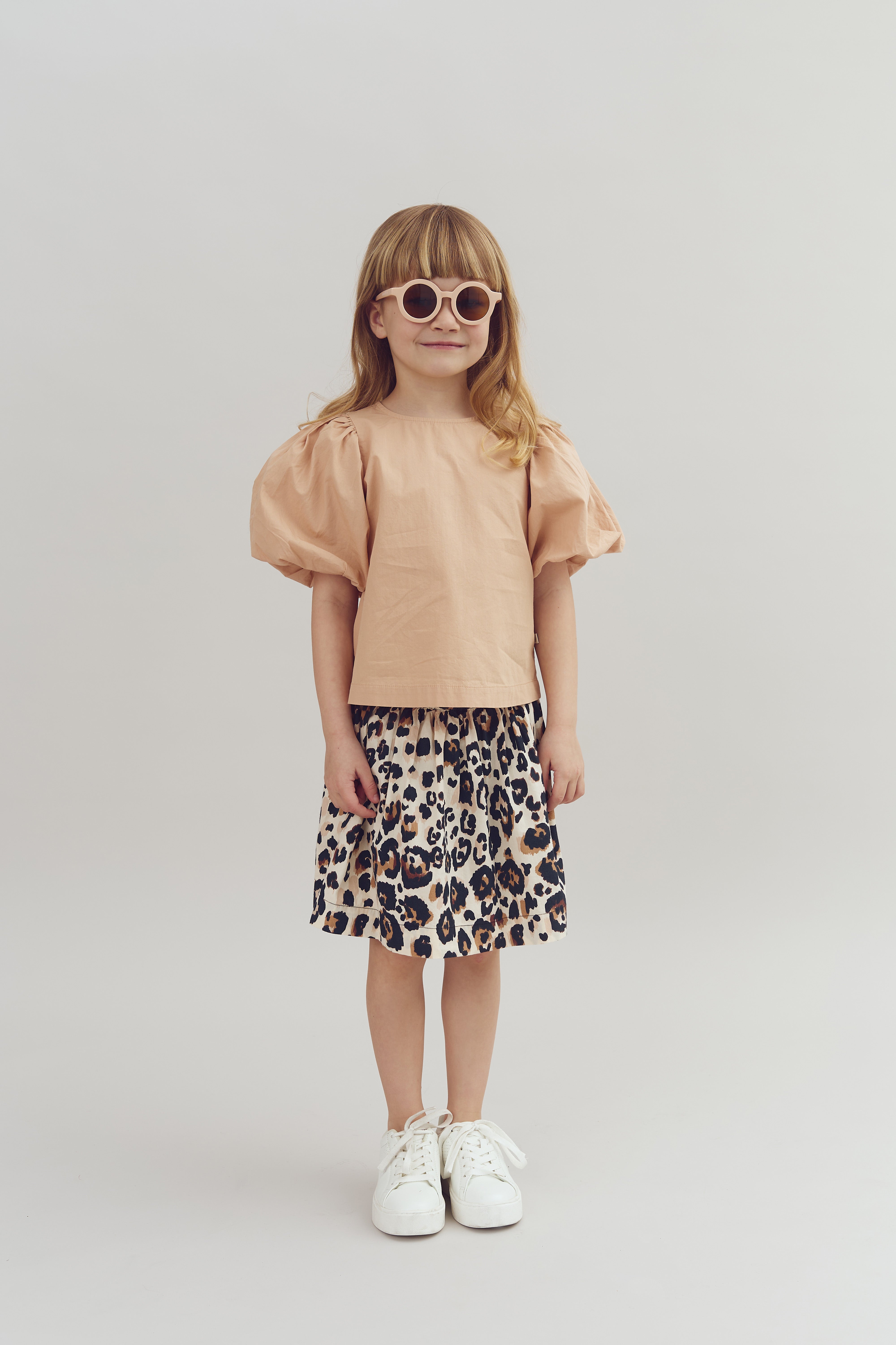 A young girl stands smiling, wearing sunglasses, a peach top with puffed sleeves, a leopard-print skirt, and white sneakers, against a plain light gray background.