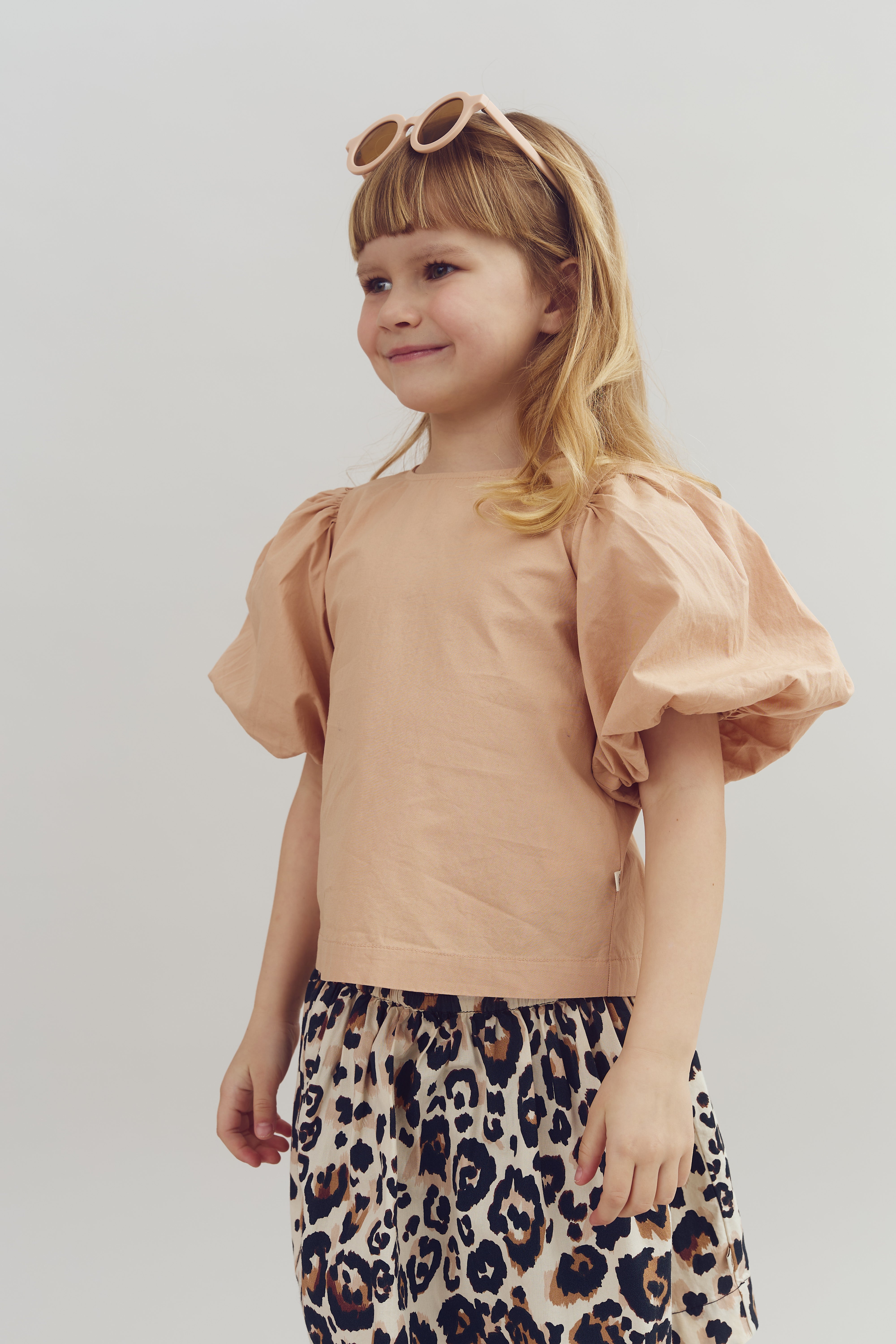 A child wears sunglasses on her head and smiles, dressed in a peach top with puffed sleeves and a leopard-print skirt, against a plain light background.