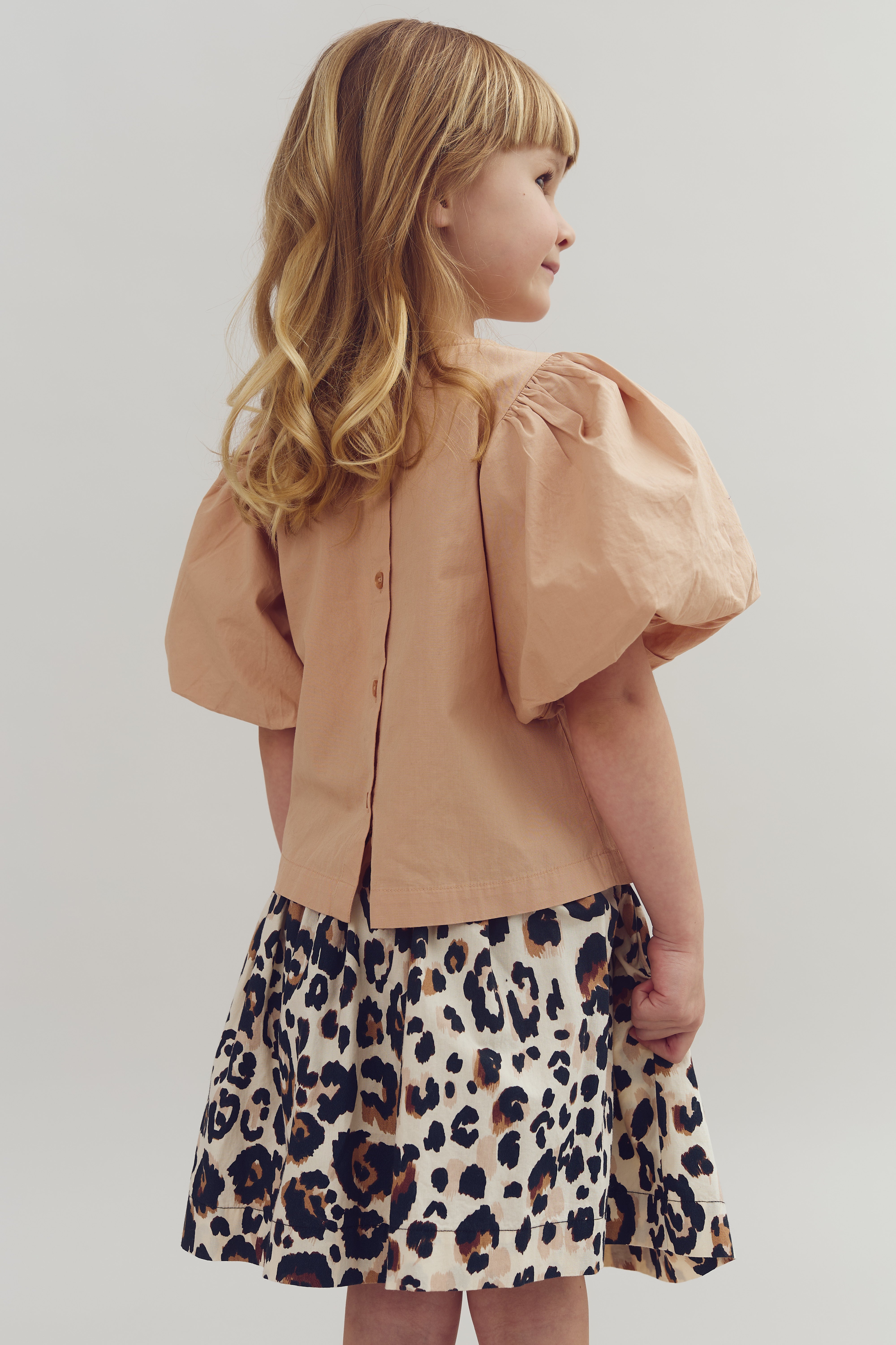 A young girl stands sideways, wearing a peach puff-sleeve shirt and a leopard print skirt, in a plain studio setting. Her long hair falls in soft curls.