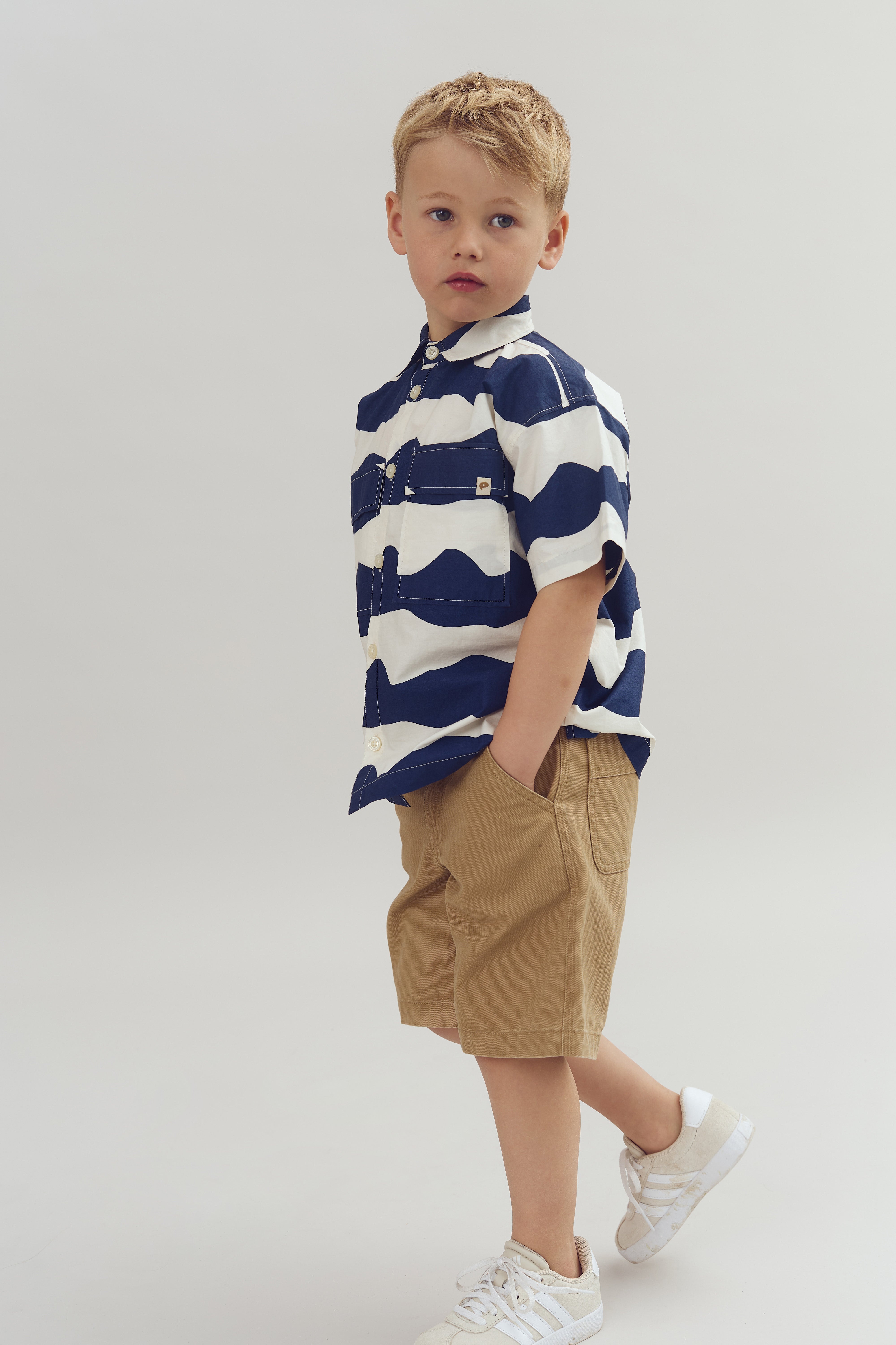 Child standing casually with hands in shorts pockets, wearing a navy and white striped shirt and white sneakers, against a plain gray background.