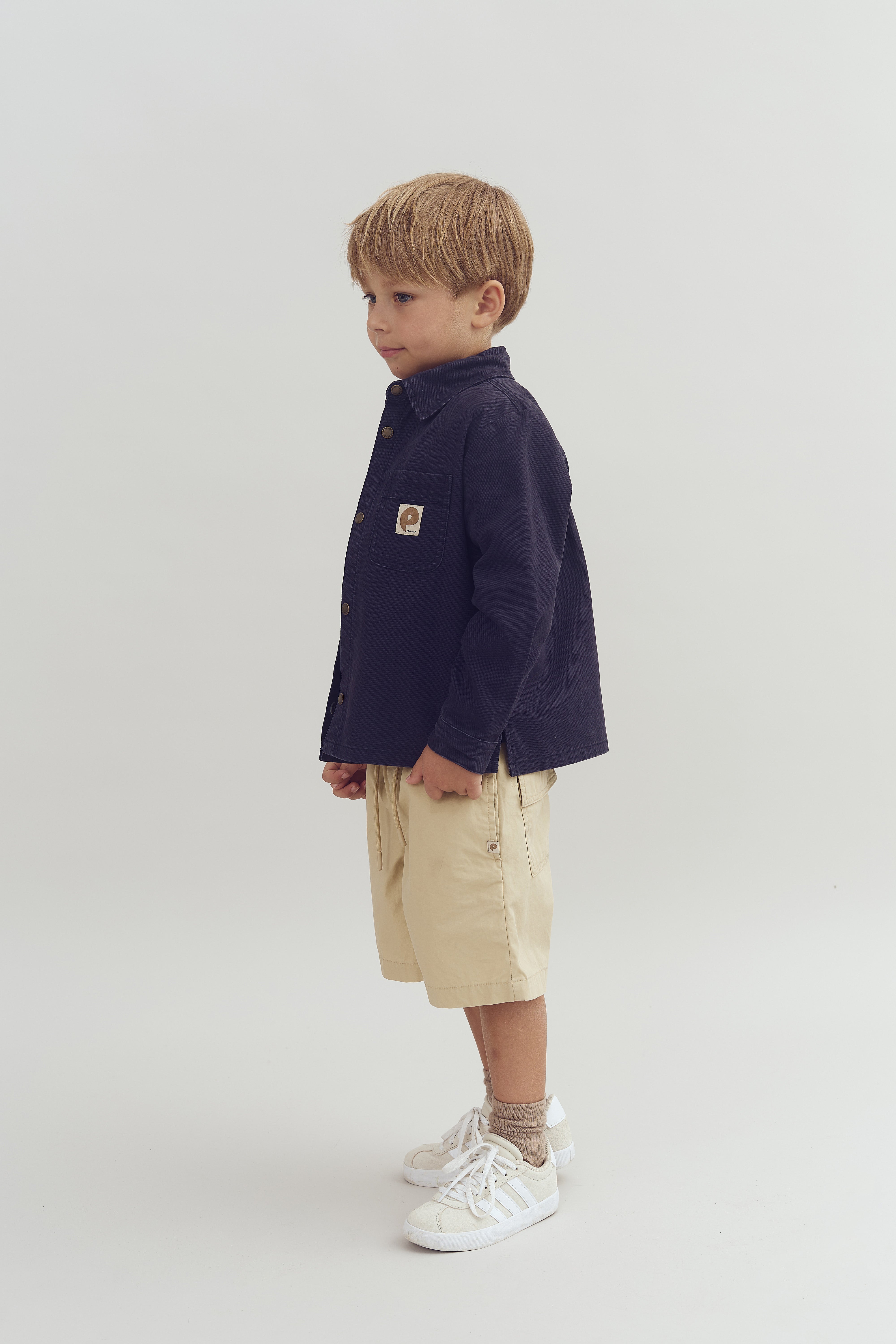 Child stands sideways wearing a navy jacket, beige shorts, and white sneakers in a neutral studio space. Jacket has a rectangular white logo patch with a stylized "P."