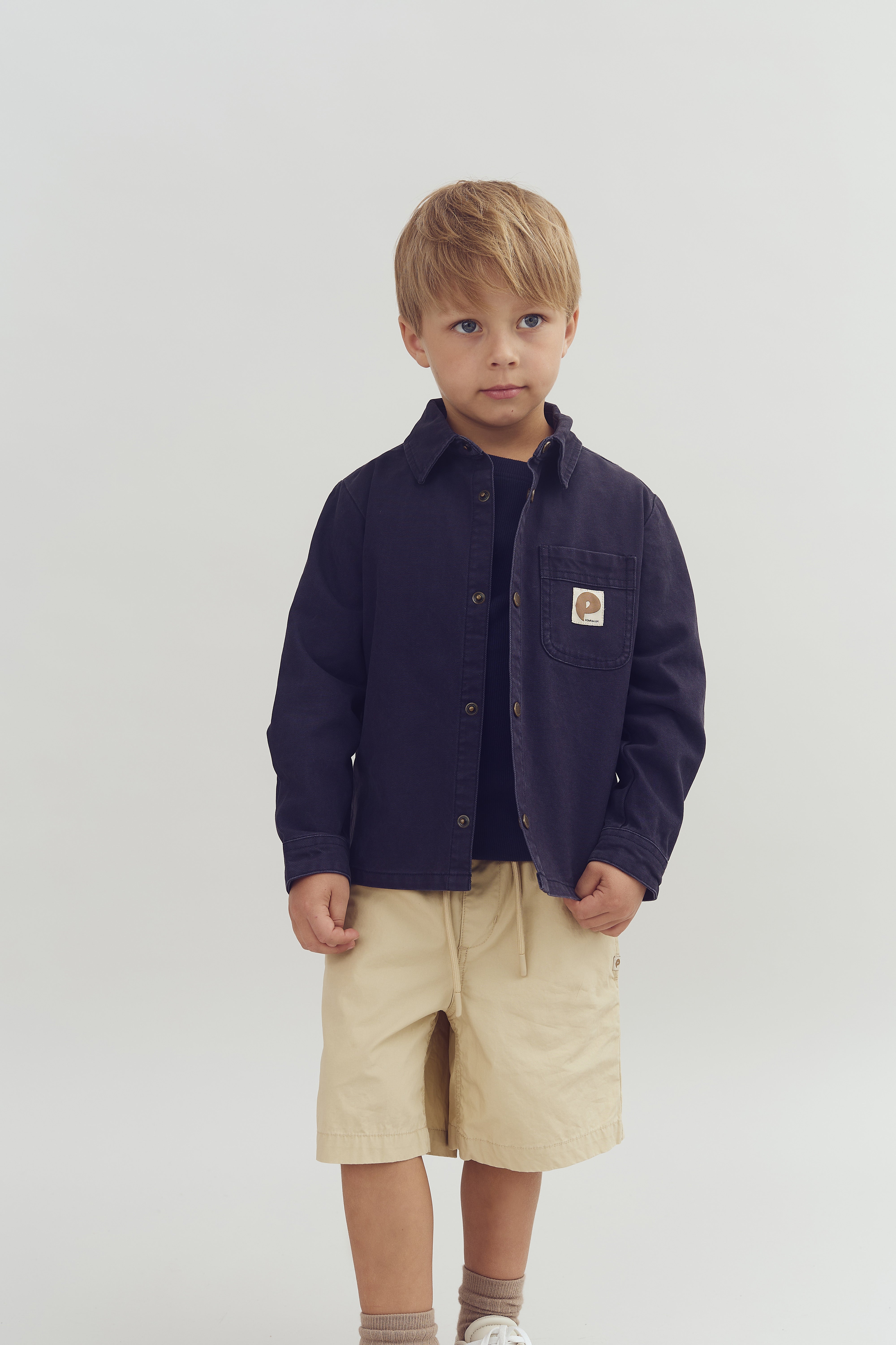 A young child stands wearing a navy jacket with a pocket logo, beige shorts, and sneakers. The background is a neutral, light-colored studio setting.