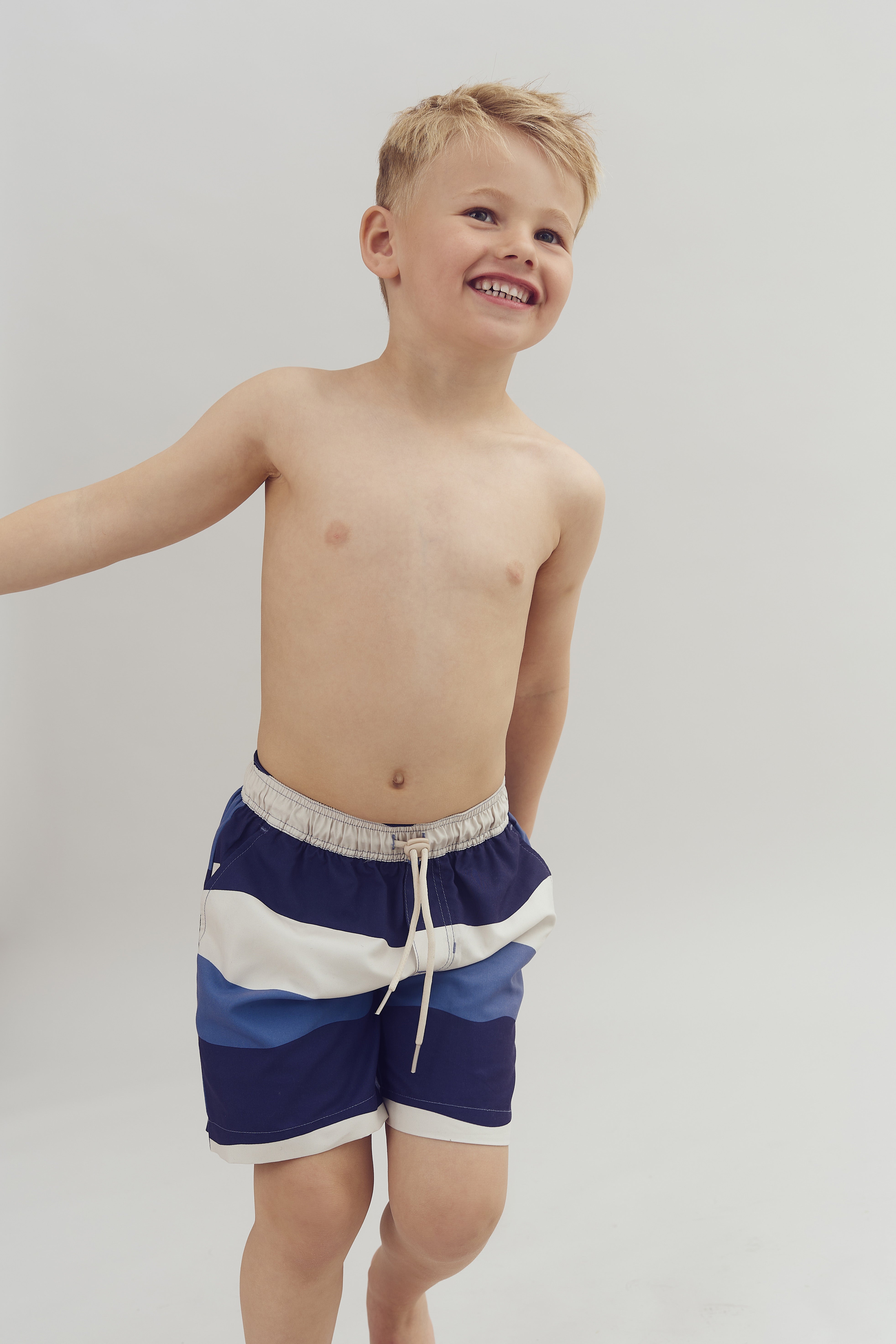 A smiling child stands shirtless, wearing blue and white shorts with a drawstring, against a plain light-colored background.