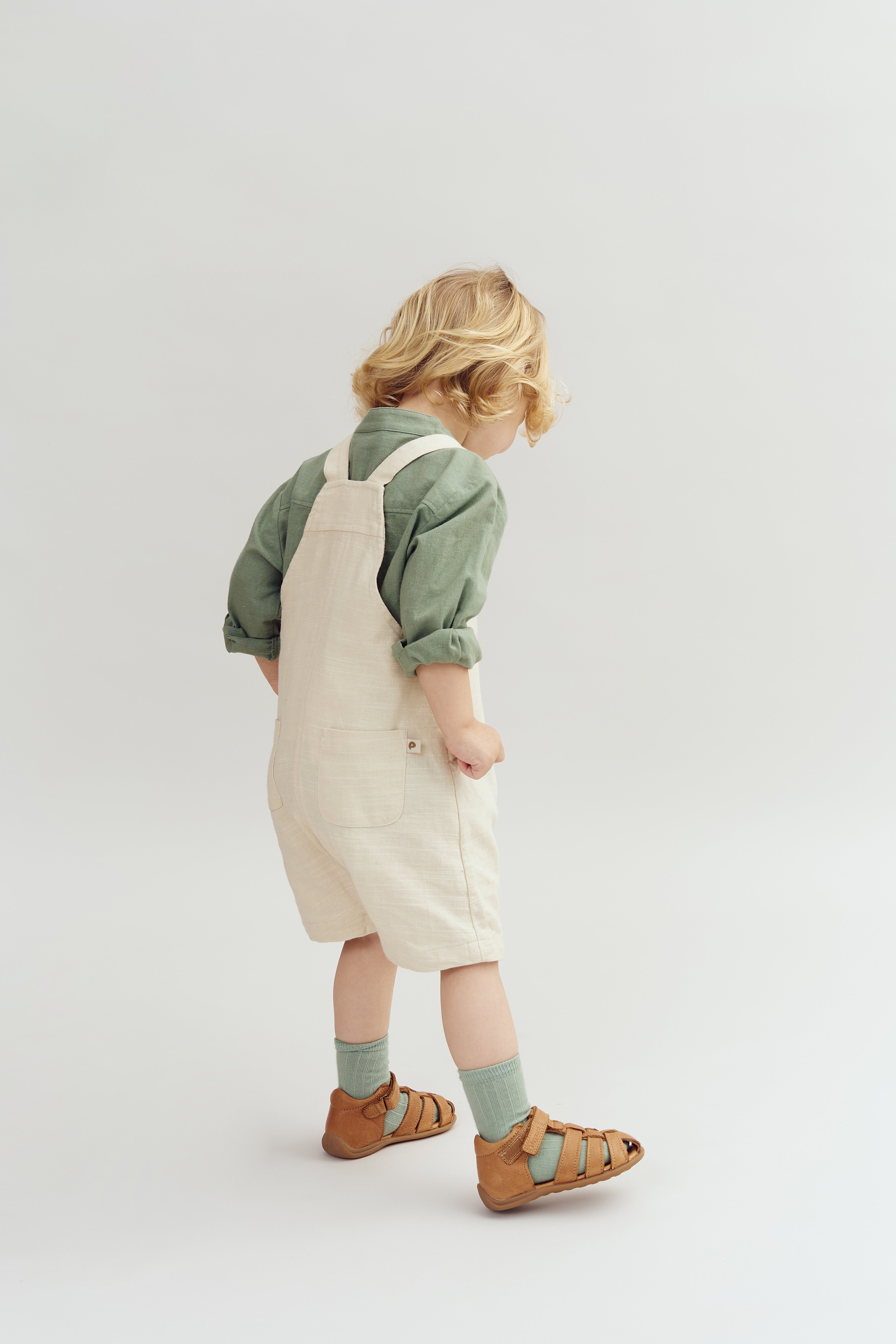 A child, wearing a green shirt and beige overalls, stands with head down. The child is also wearing brown sandals and green socks, in a minimalist, light-colored studio setting.