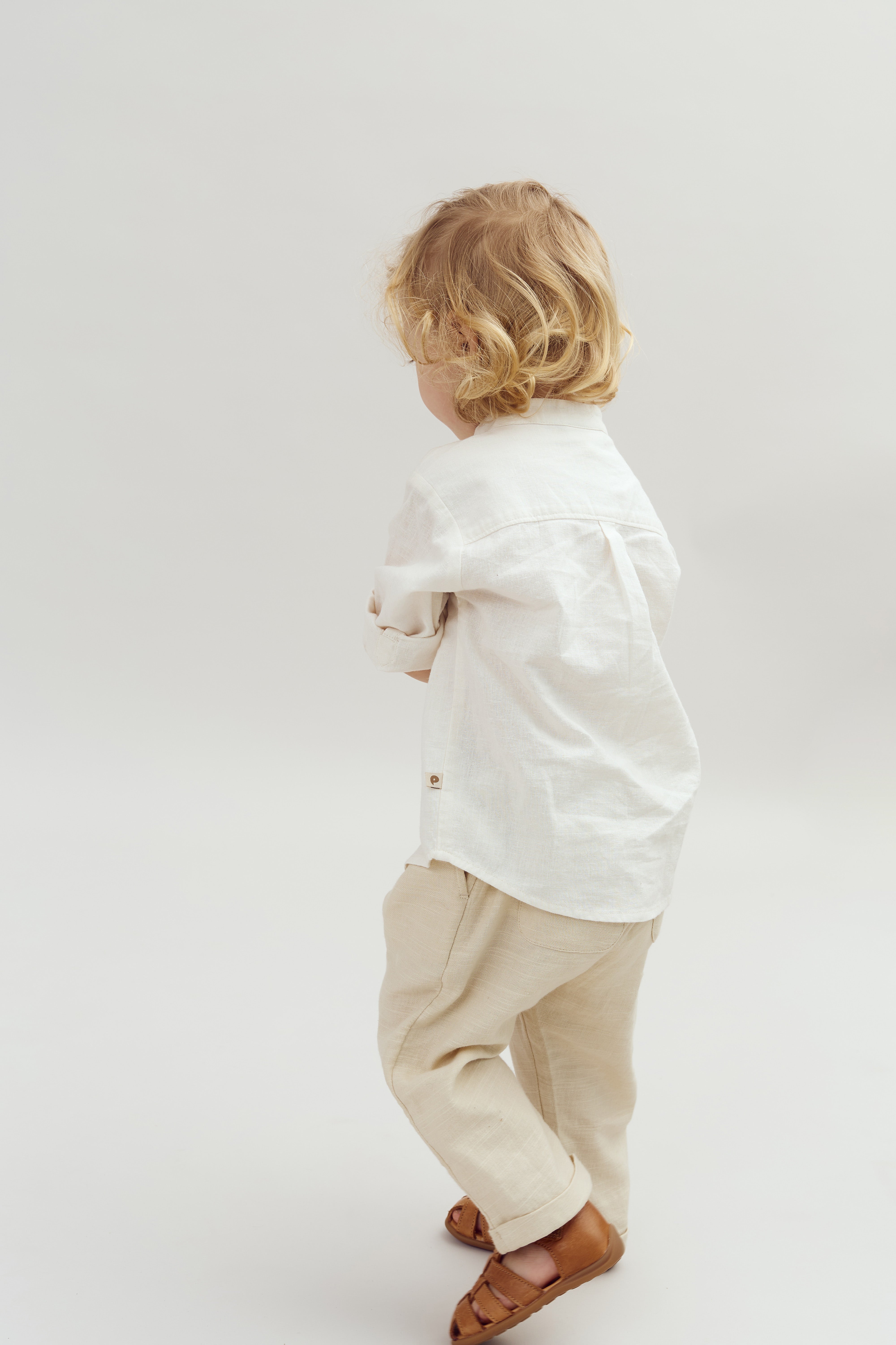 Child wearing a white shirt and beige pants walks away in a minimalistic, bright studio setting. The child has curly blonde hair and brown sandals, facing a plain background.