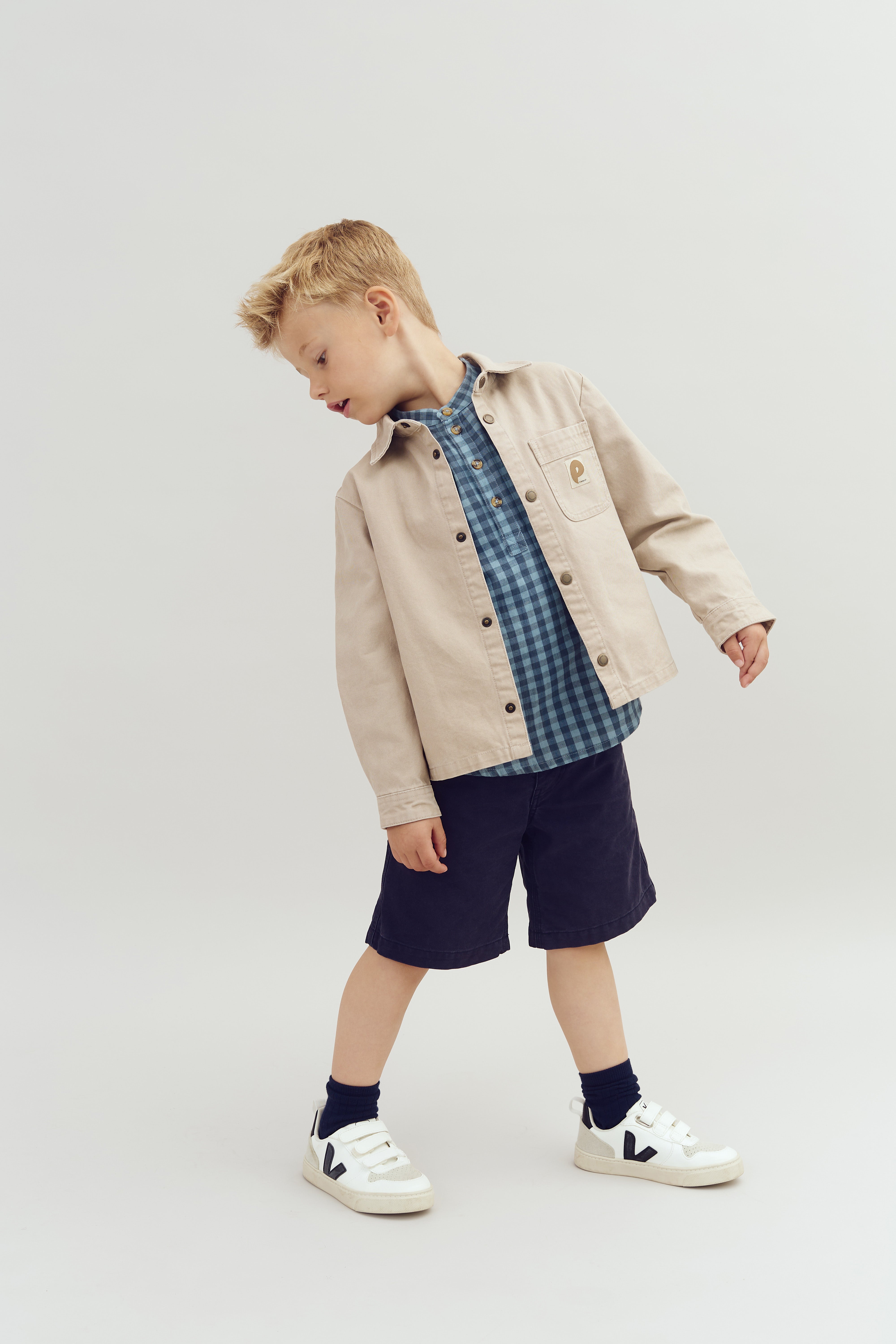 Child wearing a beige jacket and blue checkered shirt stands in a relaxed pose, looking sideways. They are in navy shorts and white sneakers on a plain, light background.