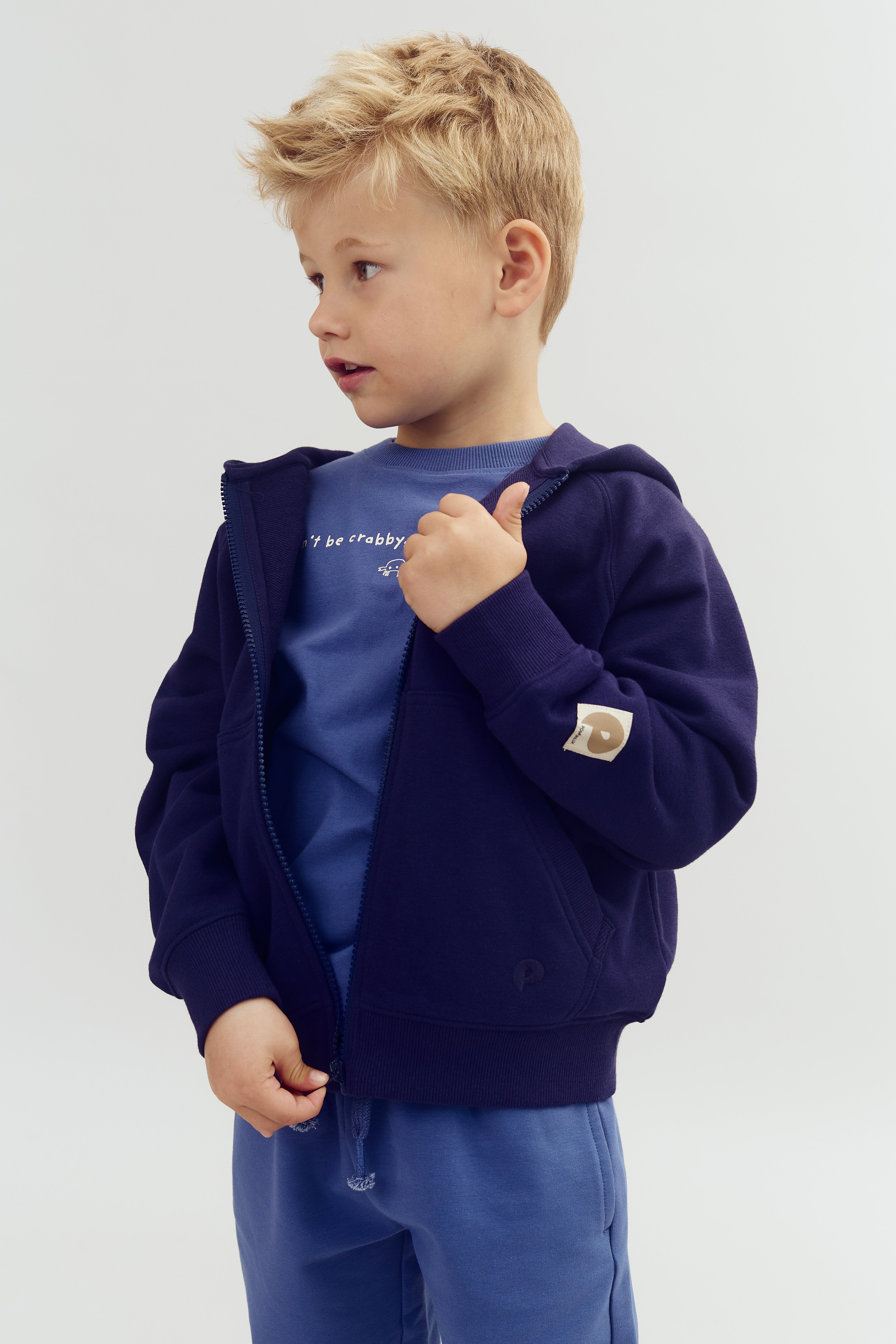 A young child wearing a navy hoodie and blue pants gestures with their thumb. The shirt reads, "Don't be crabby." The setting is a plain, light-colored background.
