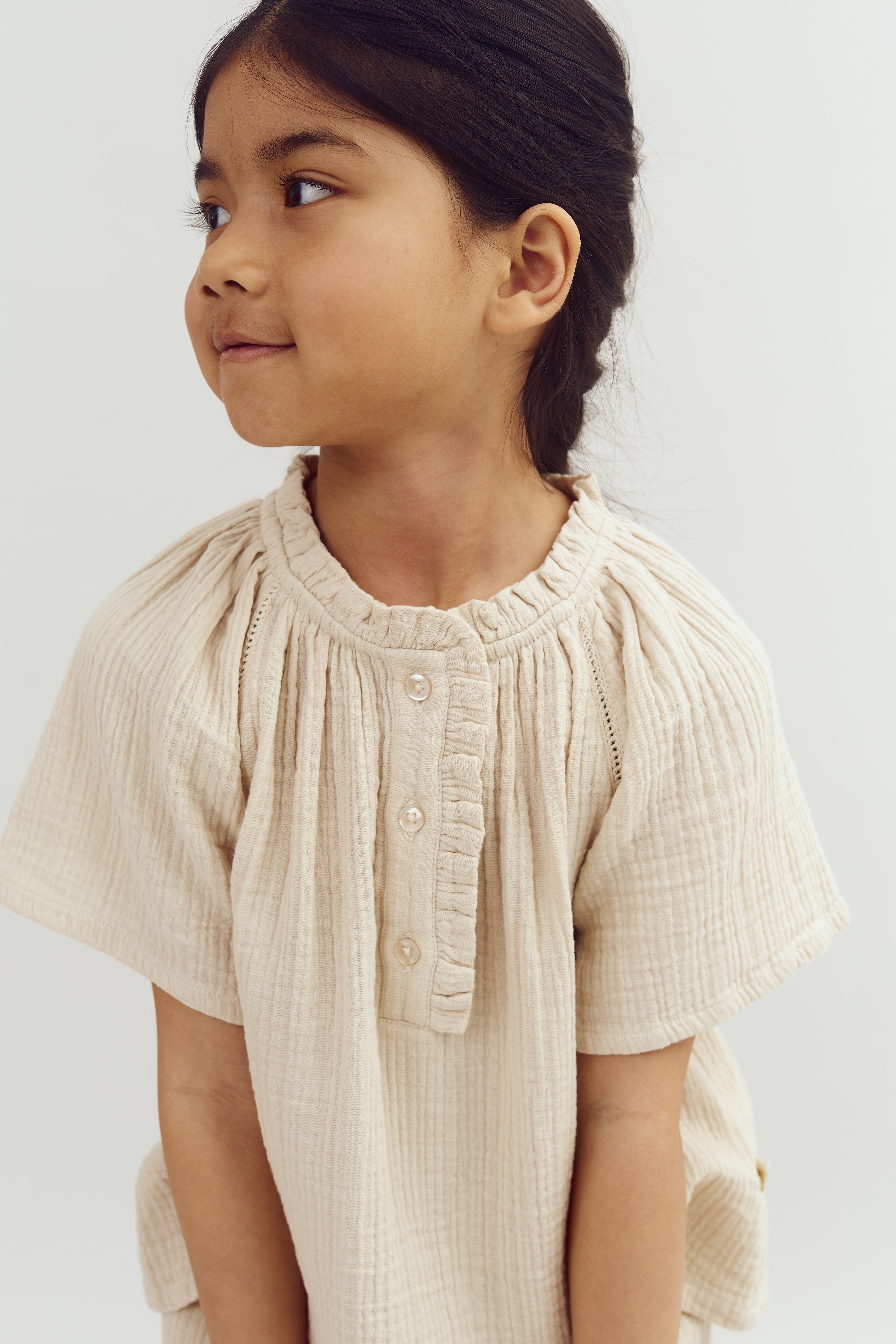 A young child with dark hair gazes to the side, wearing a light beige, textured dress with buttons. The background is plain white, emphasizing the child's profile and attire.