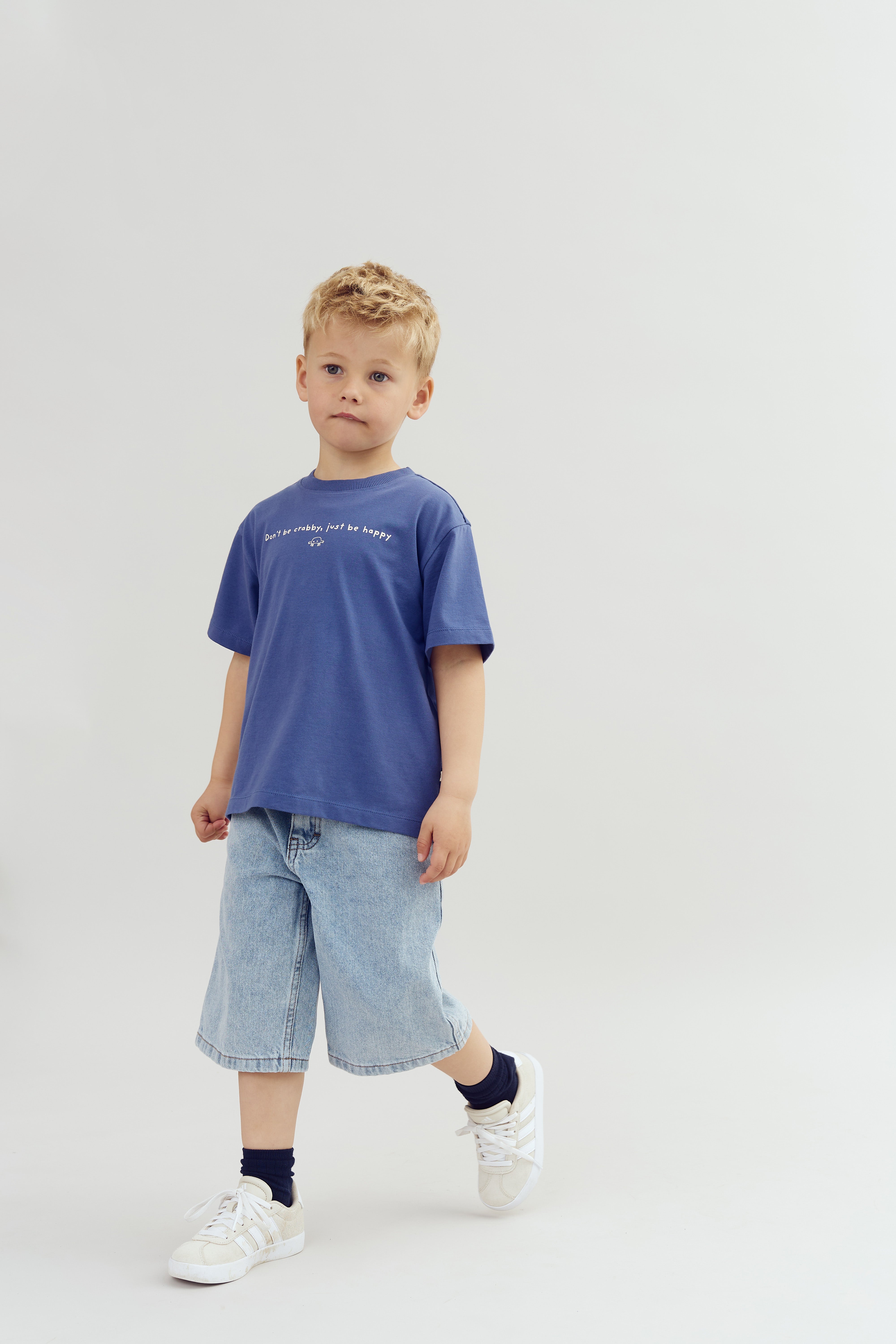 A child in a blue shirt is walking, with text "Don’t be crabby, just be happy" on the shirt, wearing denim shorts and sneakers, against a plain background.