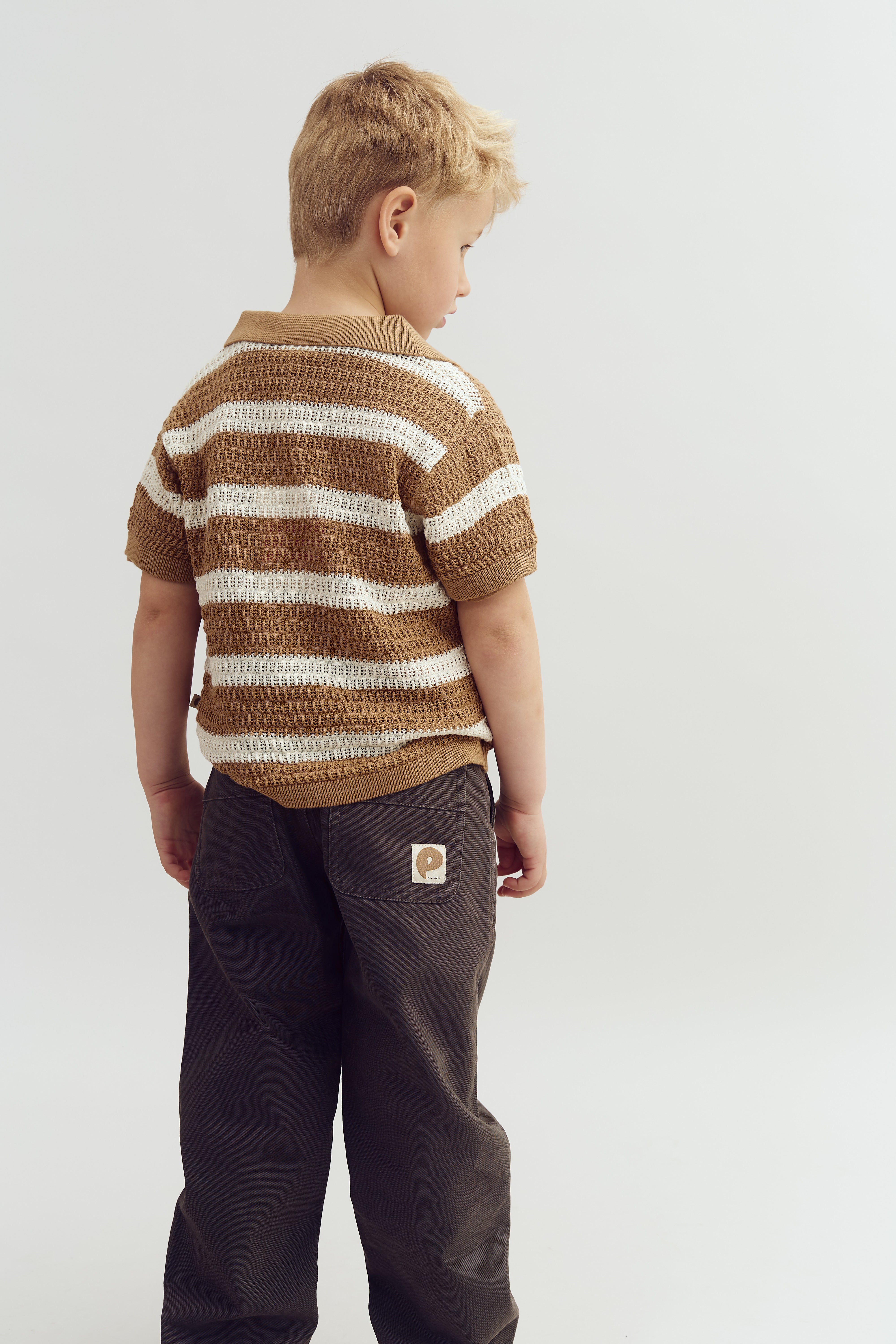 A child wearing a brown and white striped sweater and dark pants stands facing away in a neutral, light-colored setting. A pocket on the pants features a patch with a stylized "p".