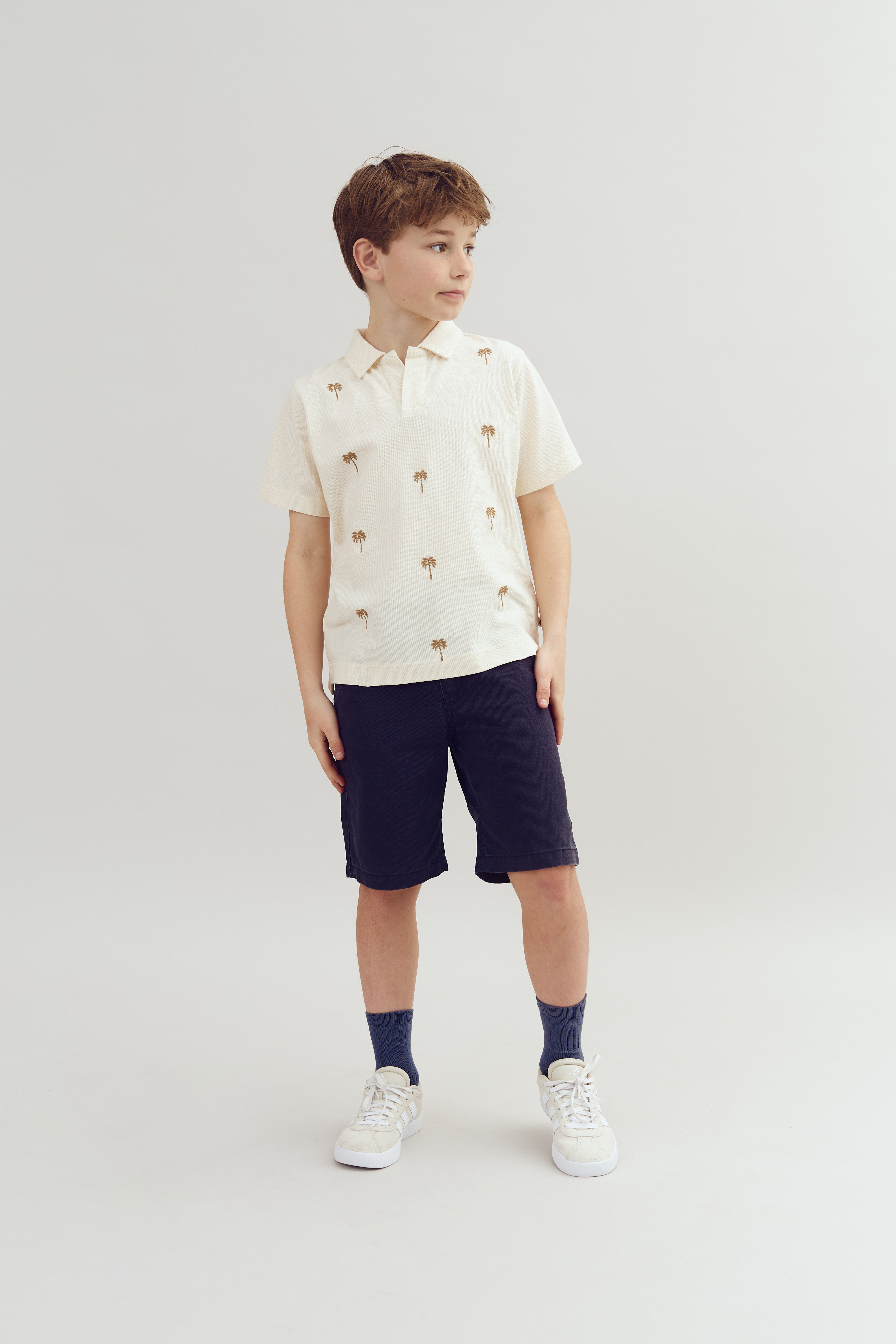 A boy stands, looking to the side, wearing a cream shirt with palm tree patterns, dark shorts, blue socks, and white sneakers, set against a plain white background.