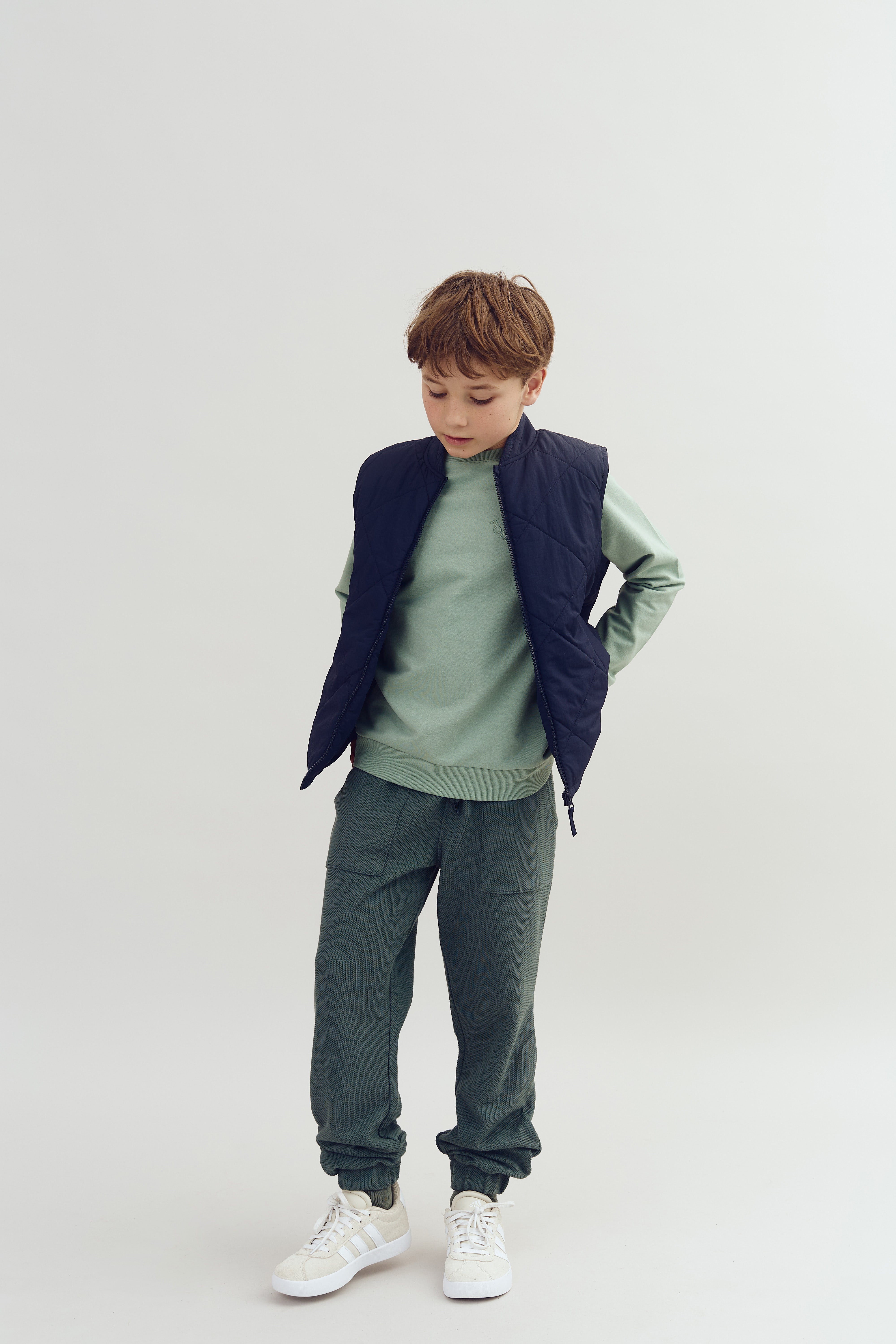 A child stands looking down, wearing a navy quilted vest, light green sweater, and green pants in a plain, light background.