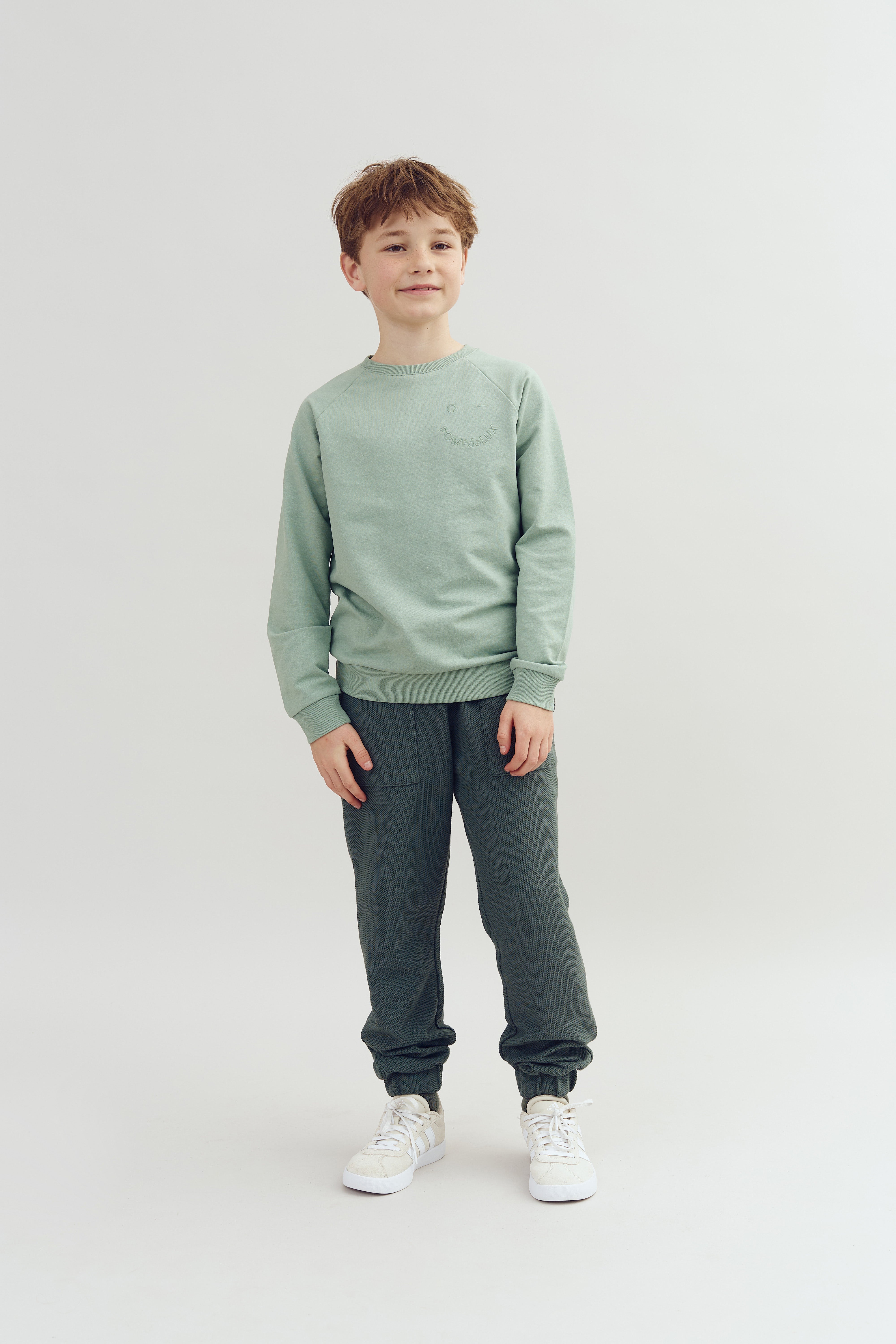 A young boy stands, smiling slightly, wearing a light green sweatshirt, dark textured pants, and white sneakers against a plain white background.
